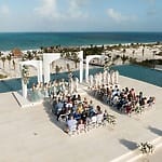 Destination wedding ceremony by the ocean with guests seated, facing the couple and wedding party.