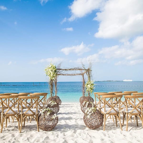 Wooden chairs and a floral arch arranged for a destination wedding on a sandy beach by the ocean.