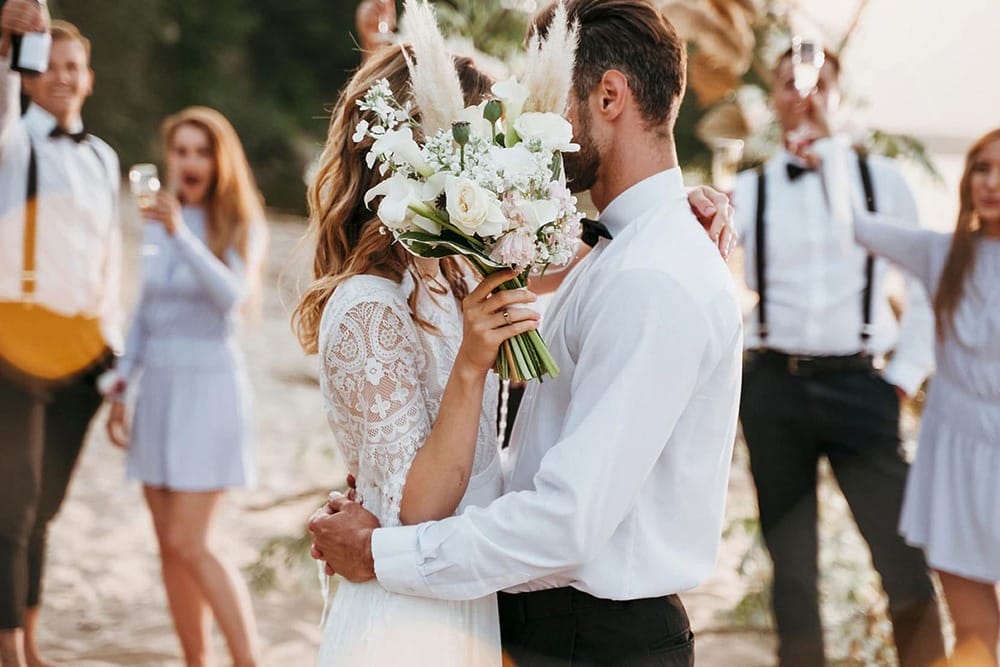 Bride and groom embrace on a beach during a destination wedding, as guests in formal attire celebrate nearby.