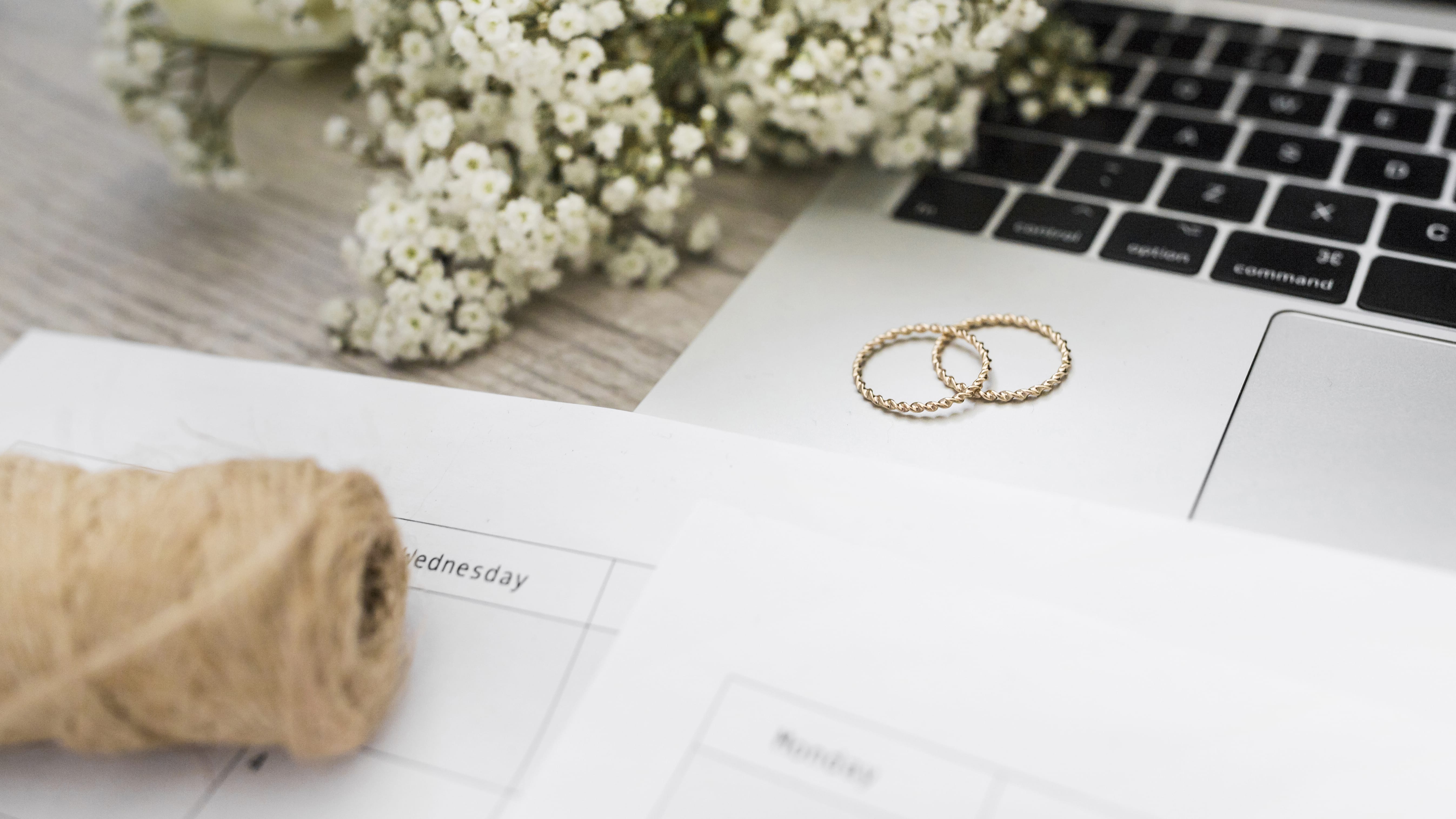 Two gold wedding rings on a laptop, next to white flowers, string, and destination wedding calendars.