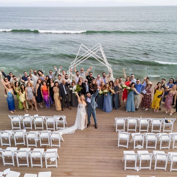 A destination wedding group stands on a deck by the ocean, empty chairs visible in the foreground.