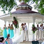 At a destination wedding, the bride and groom kiss under a gazebo while guests and the wedding party watch.