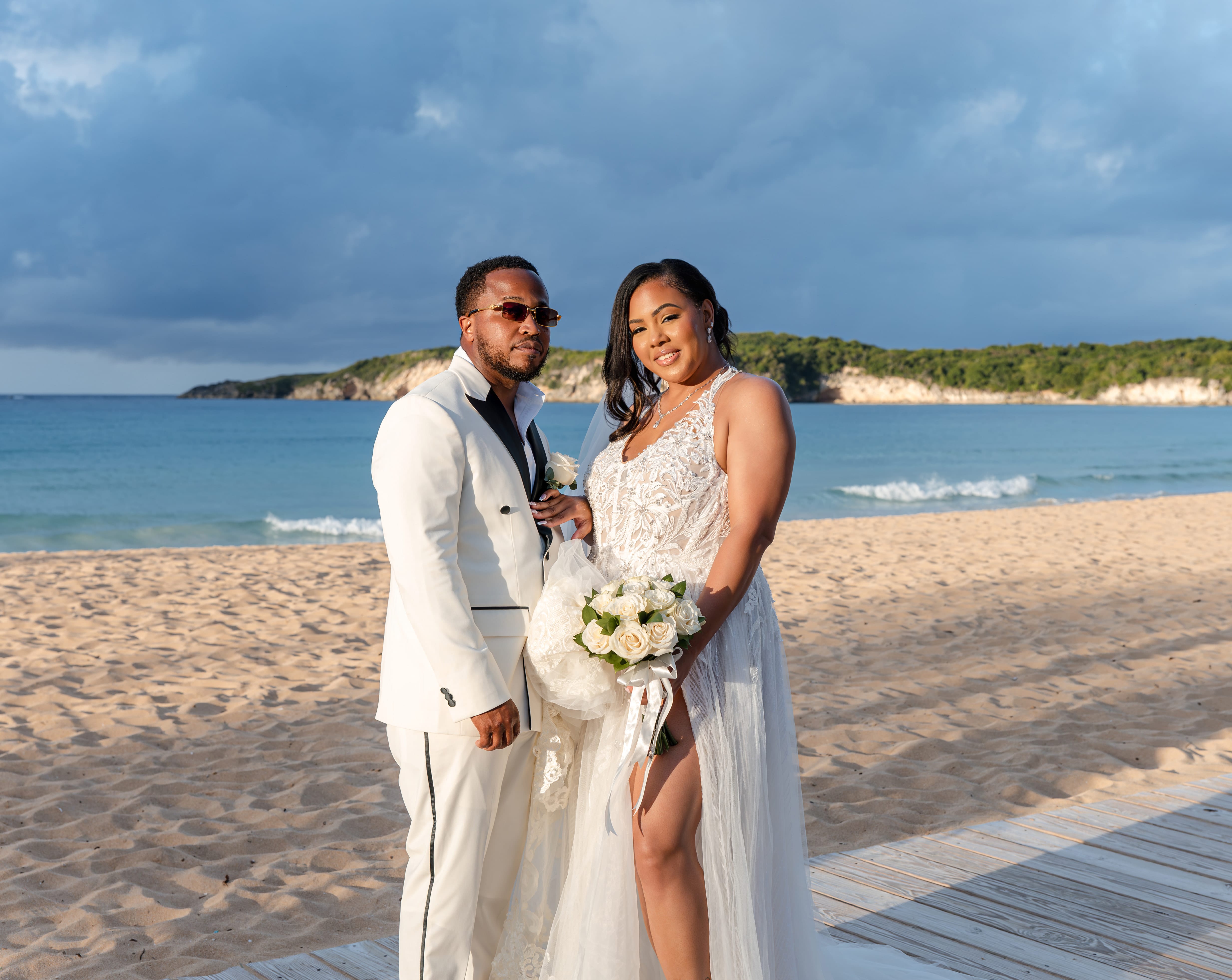 A couple in wedding attire poses on a sandy beach, ocean and cliffs framing their destination wedding.