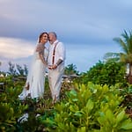 Bride and groom pose at their destination wedding outdoors, framed by lush greenery and palm trees.