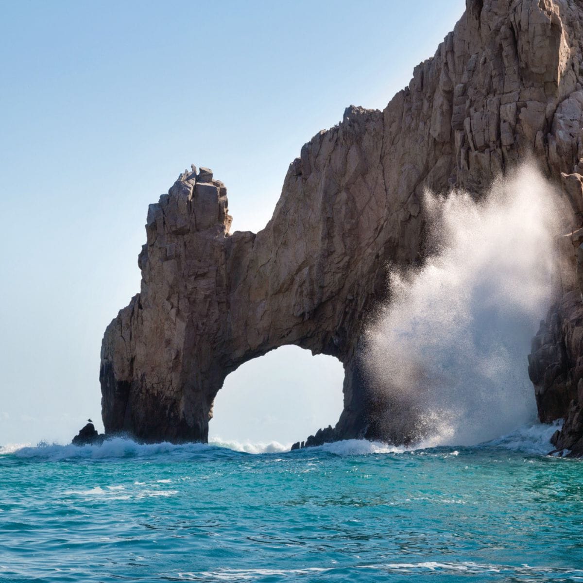 Waves crash by a rocky arch and turquoise sea, a stunning backdrop for a destination wedding.