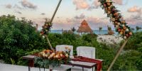 Destination wedding table featuring elegant floral centerpieces, set by the ocean with palm-thatched huts.