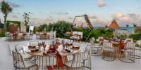 Destination wedding setup with round tables, chairs, and elegant d&eacute;cor facing the ocean at sunset.
