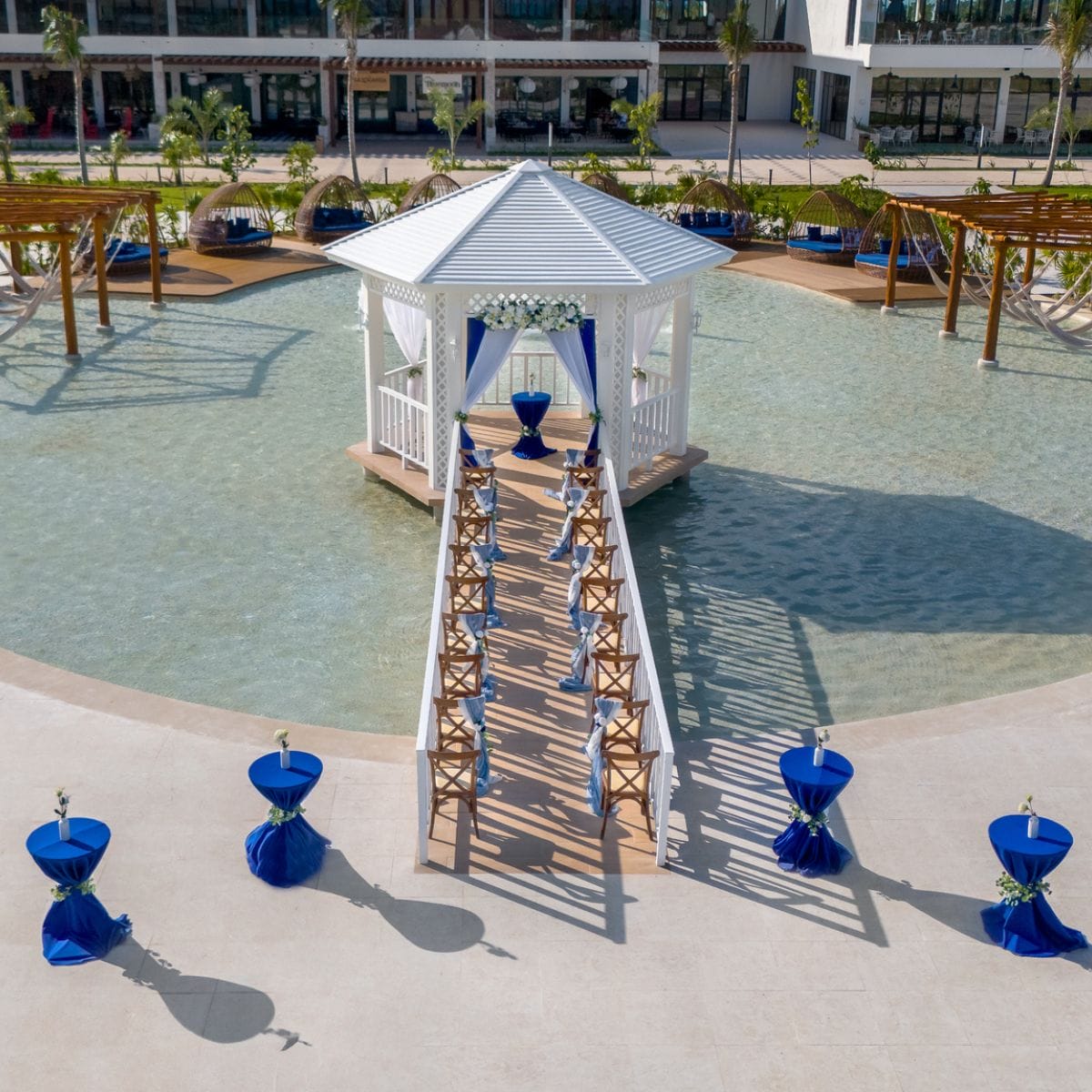 White gazebo on water with wooden aisle, set up for a destination wedding with blue-themed tables.