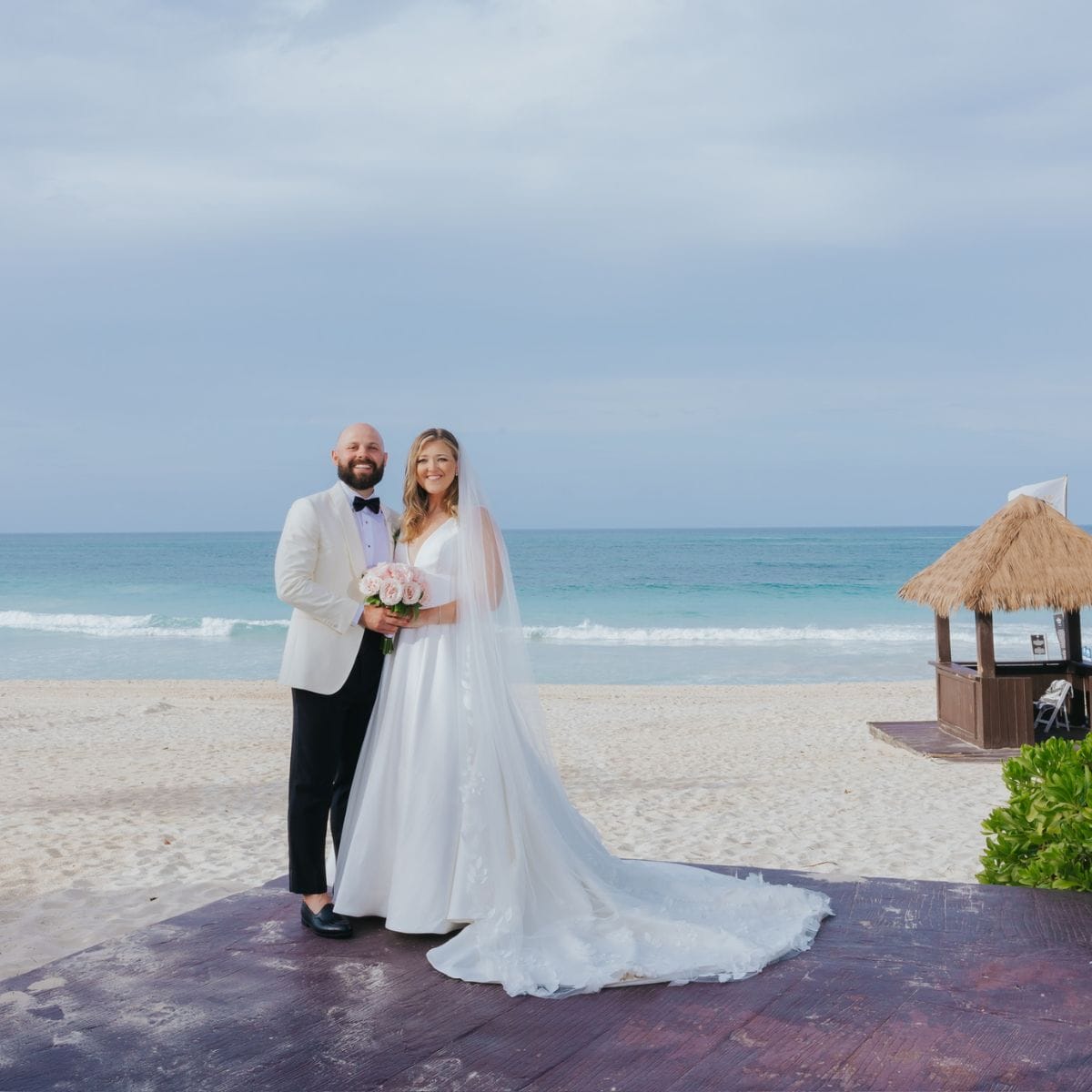A bride and groom in wedding attire stand on a beach, holding a bouquet at their destination wedding.