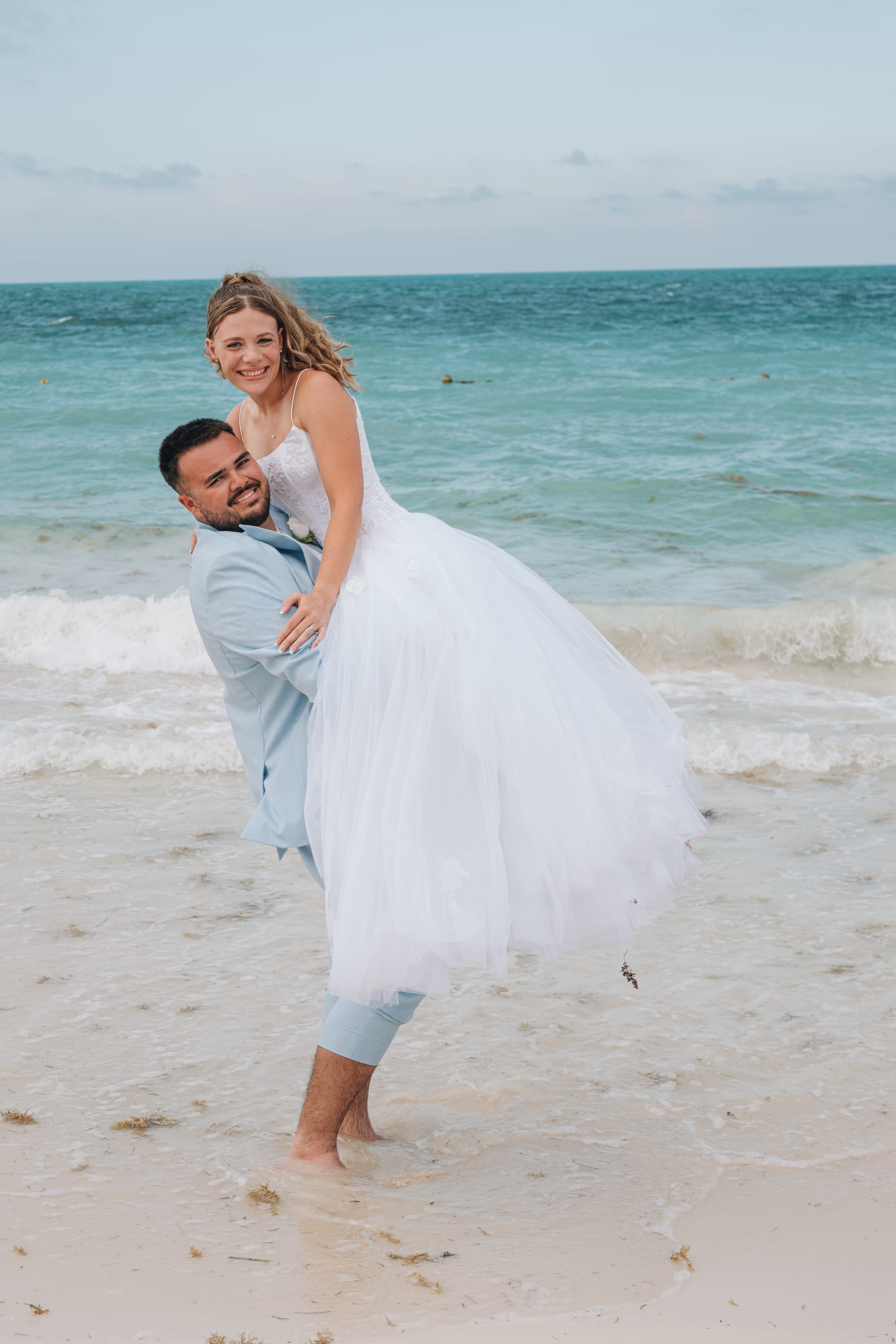 Man lifts woman in white dress on beach with waves, romantic destination wedding scene.