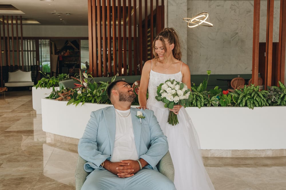 Bride with bouquet smiles beside seated groom in modern indoor destination wedding setting.