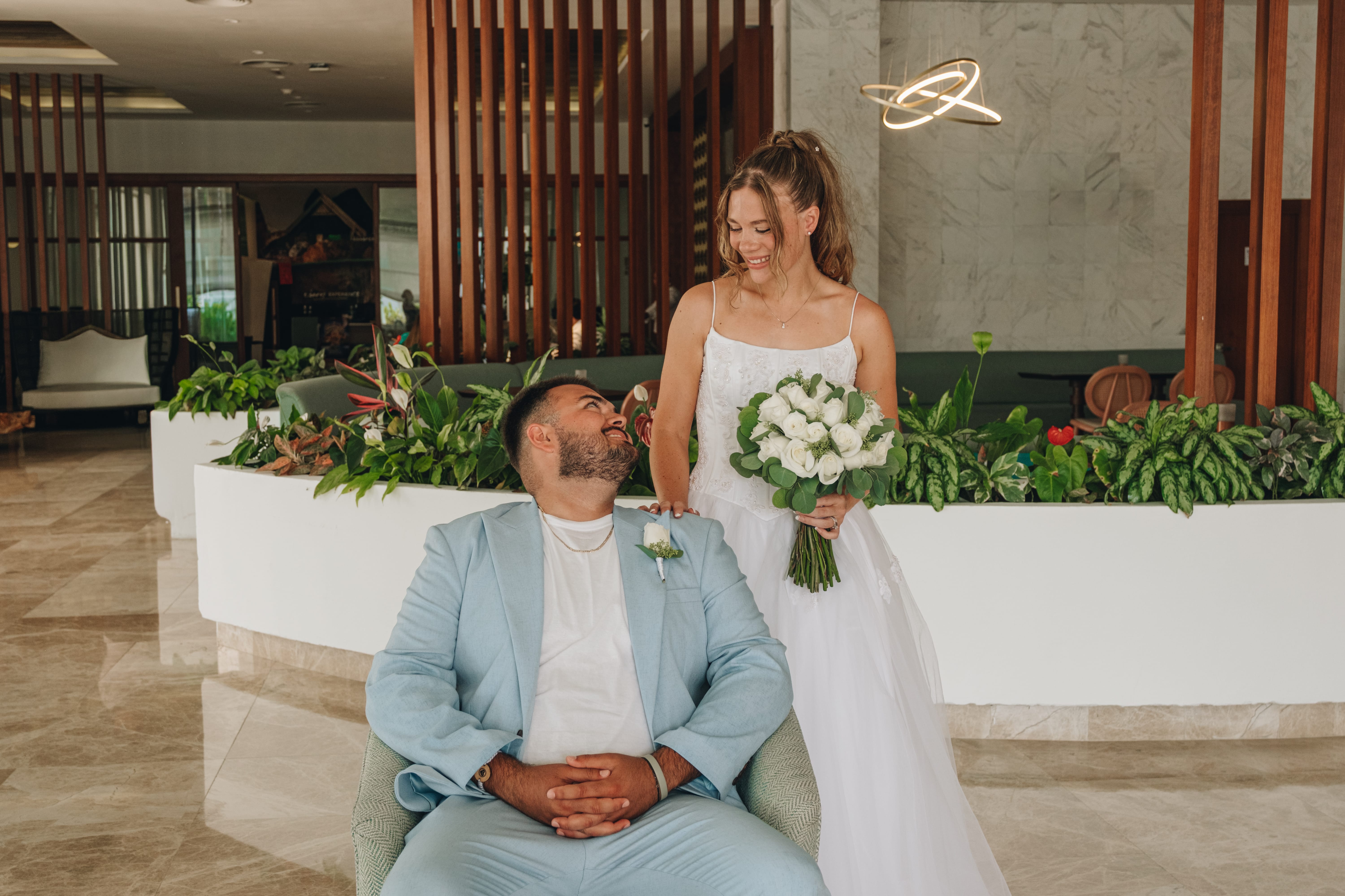 Bride with bouquet smiles beside seated groom in modern indoor destination wedding setting.