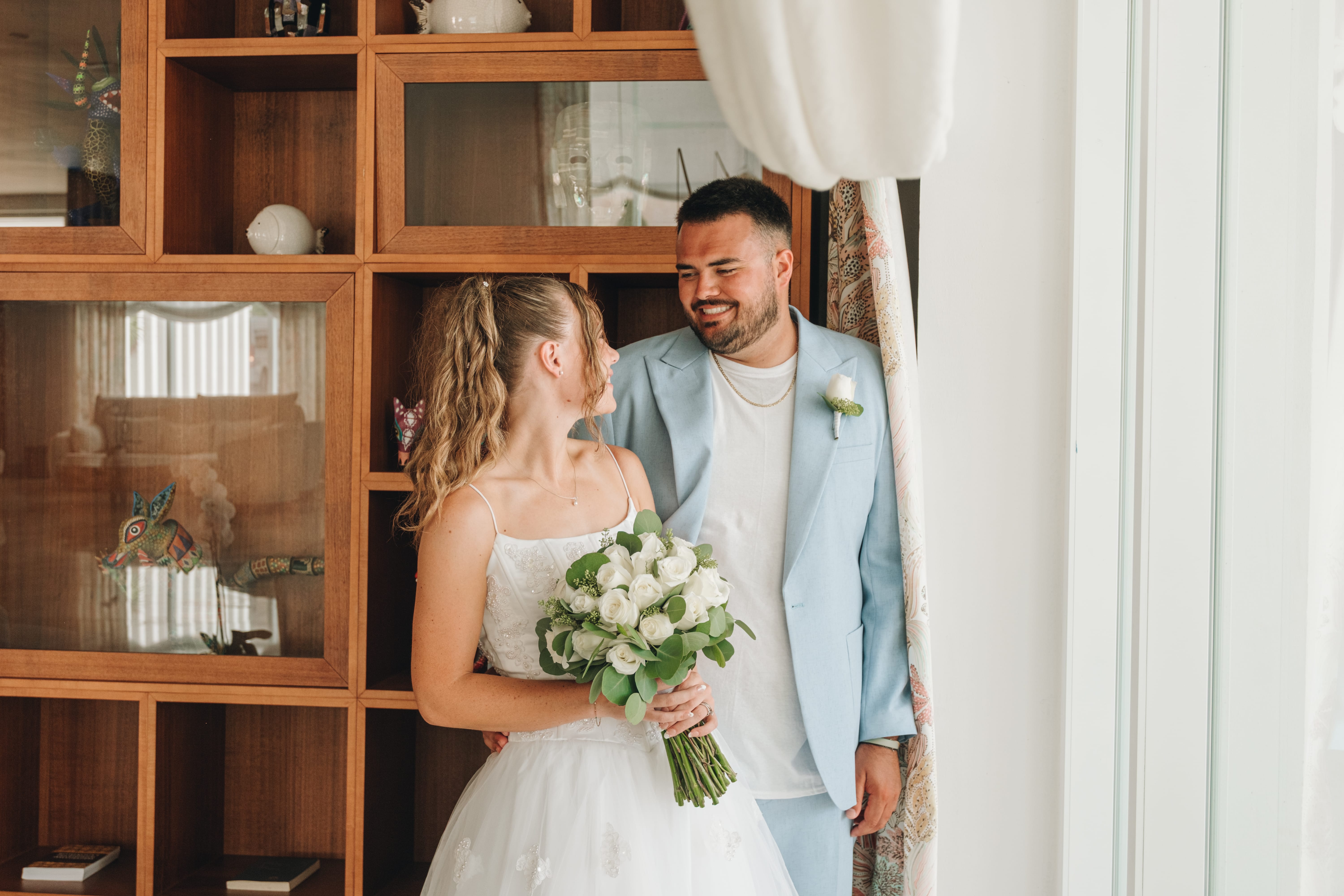 Bride with bouquet and groom in blue suit smile by shelves at their destination wedding.