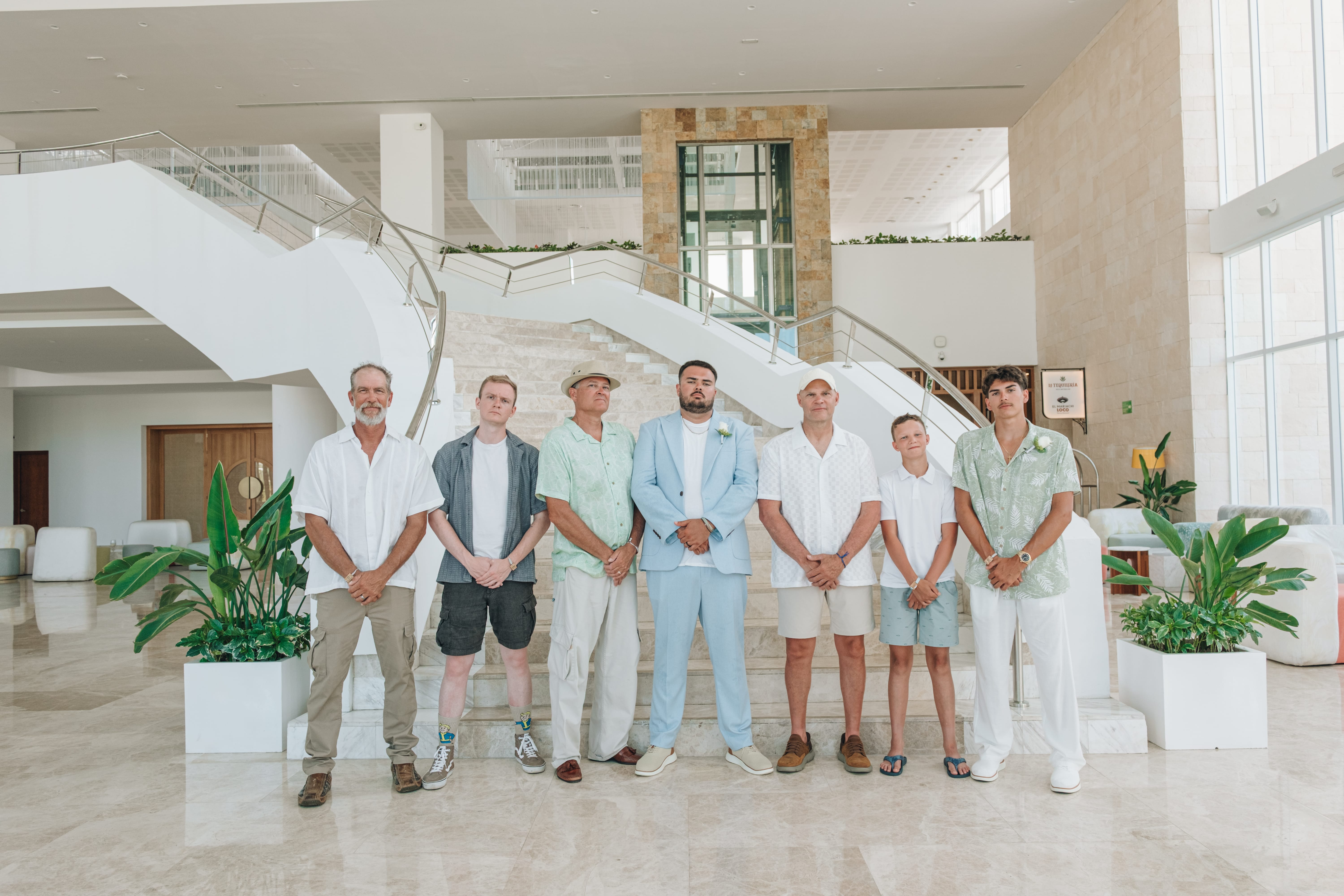 Seven men by a staircase in light, summery outfits, dressed for a destination wedding indoors.