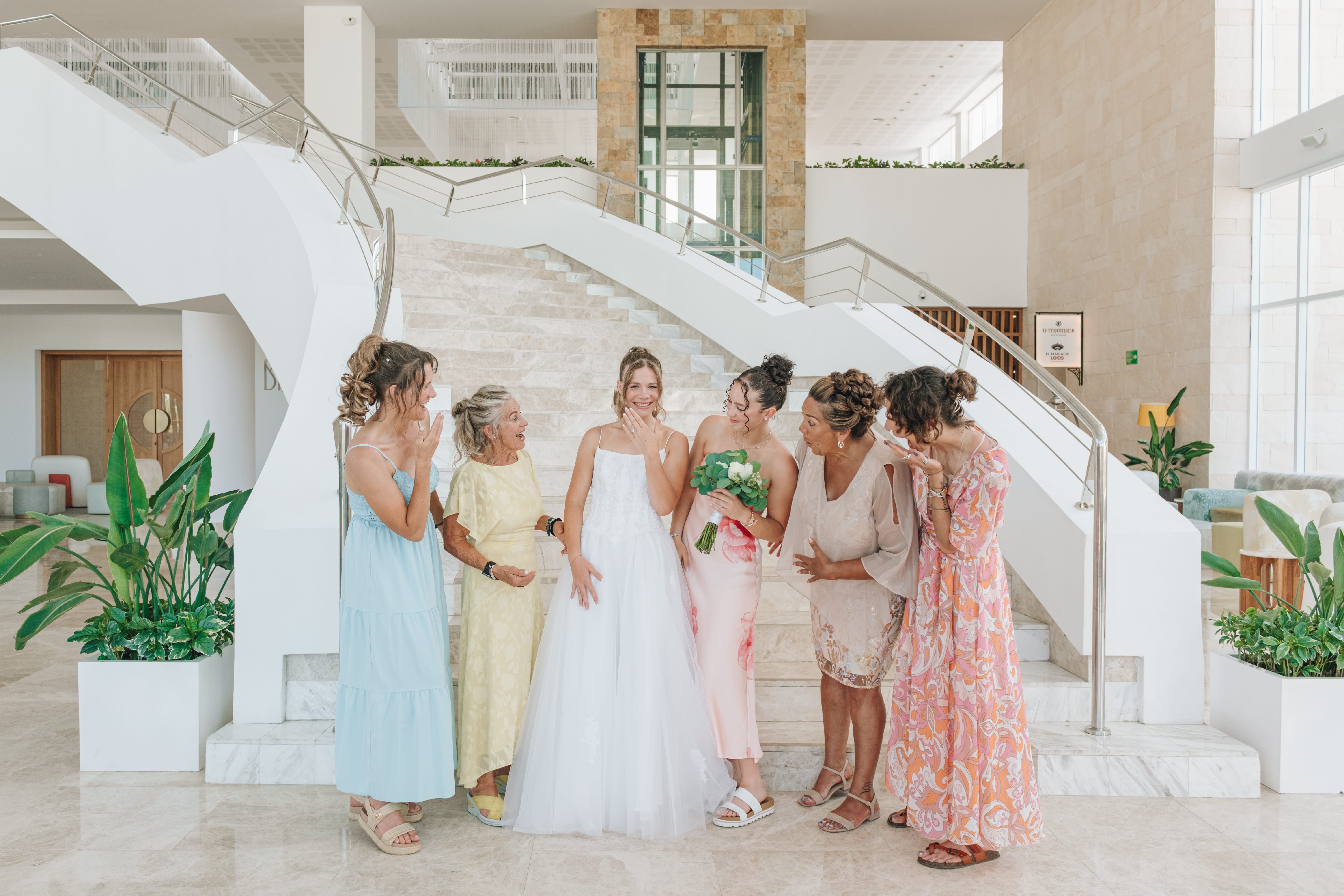 Bride in white dress with six women, all smiling at destination wedding in bright lobby with stairs.