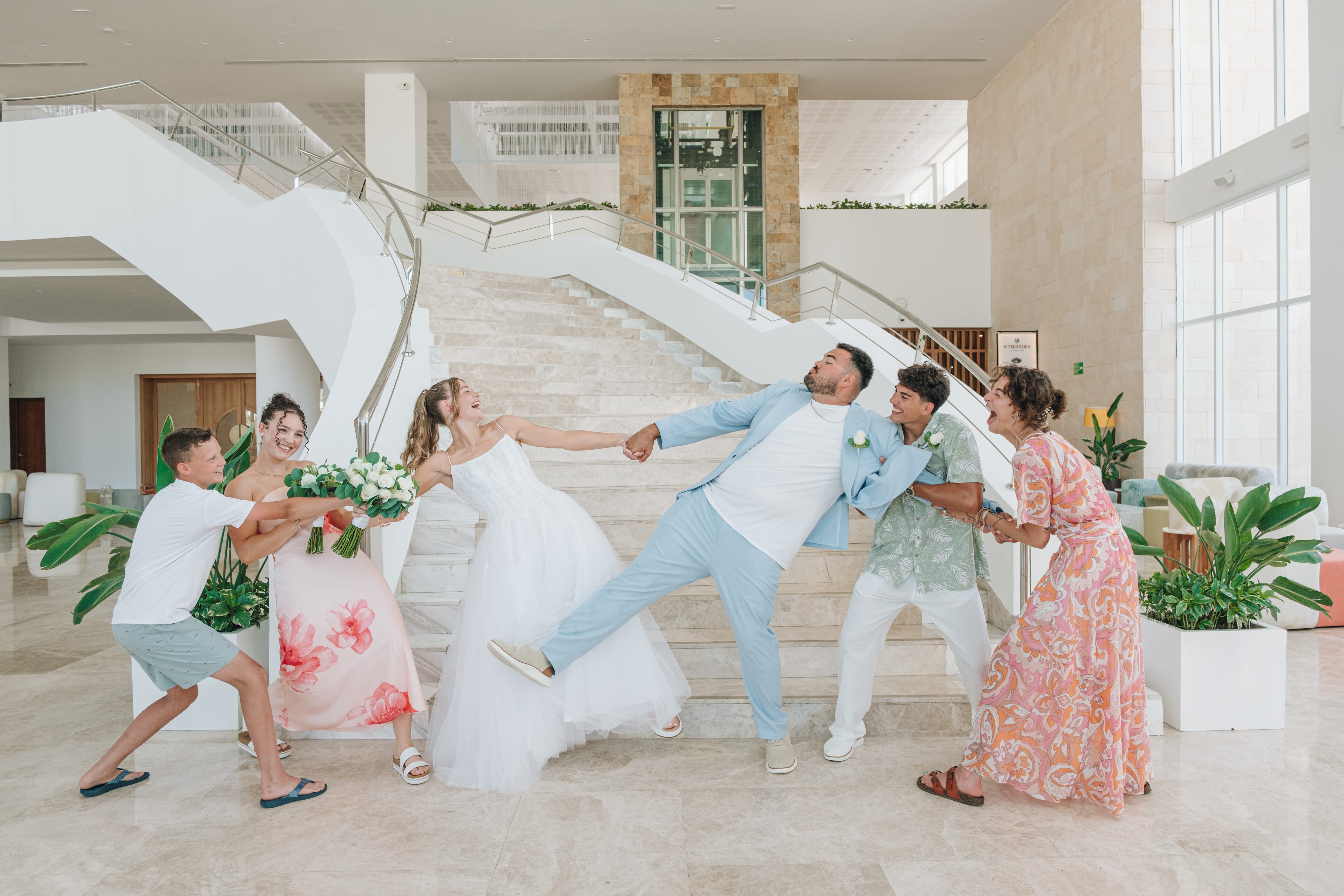 At a destination wedding, the party playfully tugs Kayleigh and Isaac on an indoor staircase.