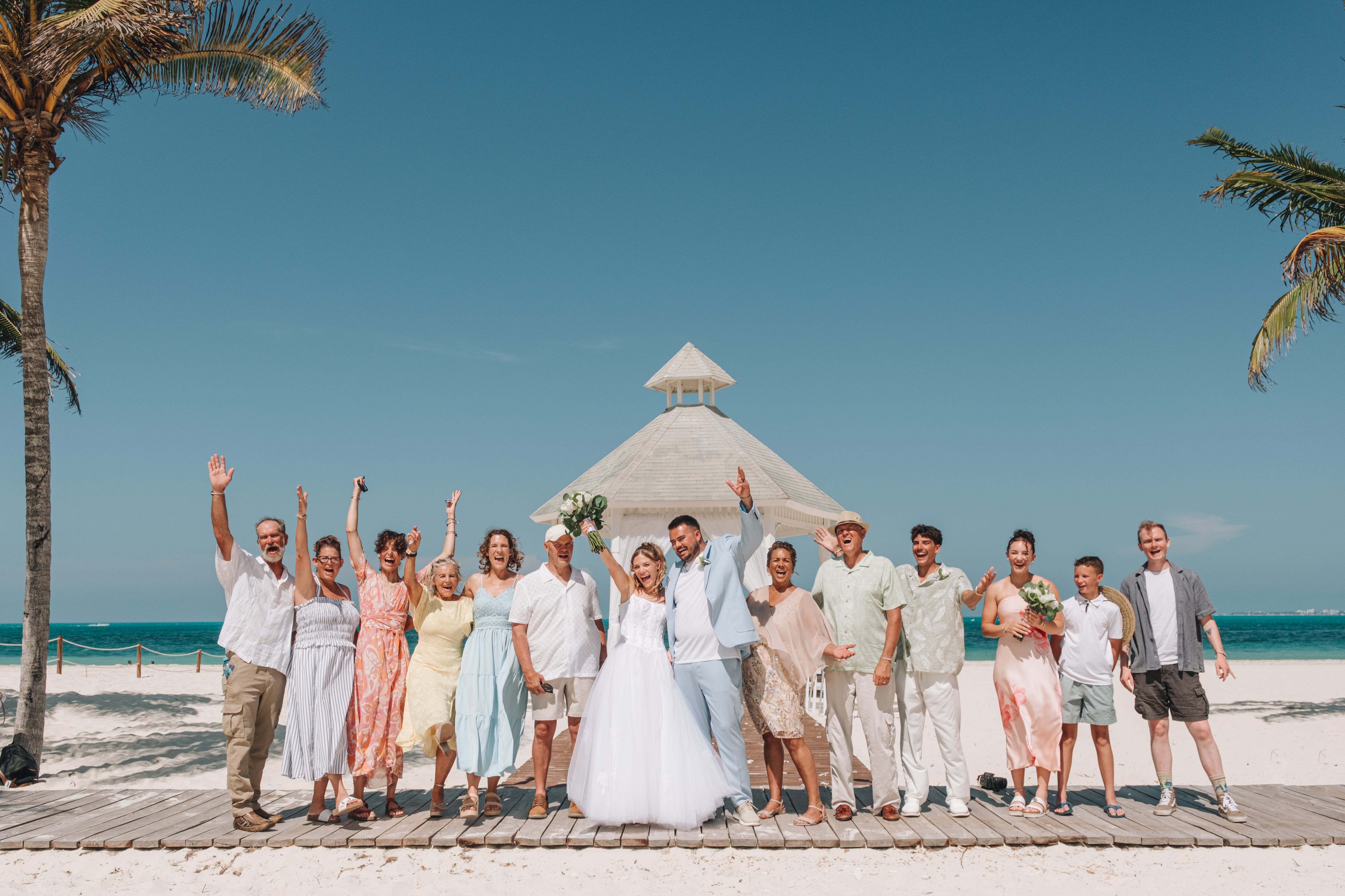 The wedding party poses on a sunny beach with palm trees and a gazebo at a destination wedding.