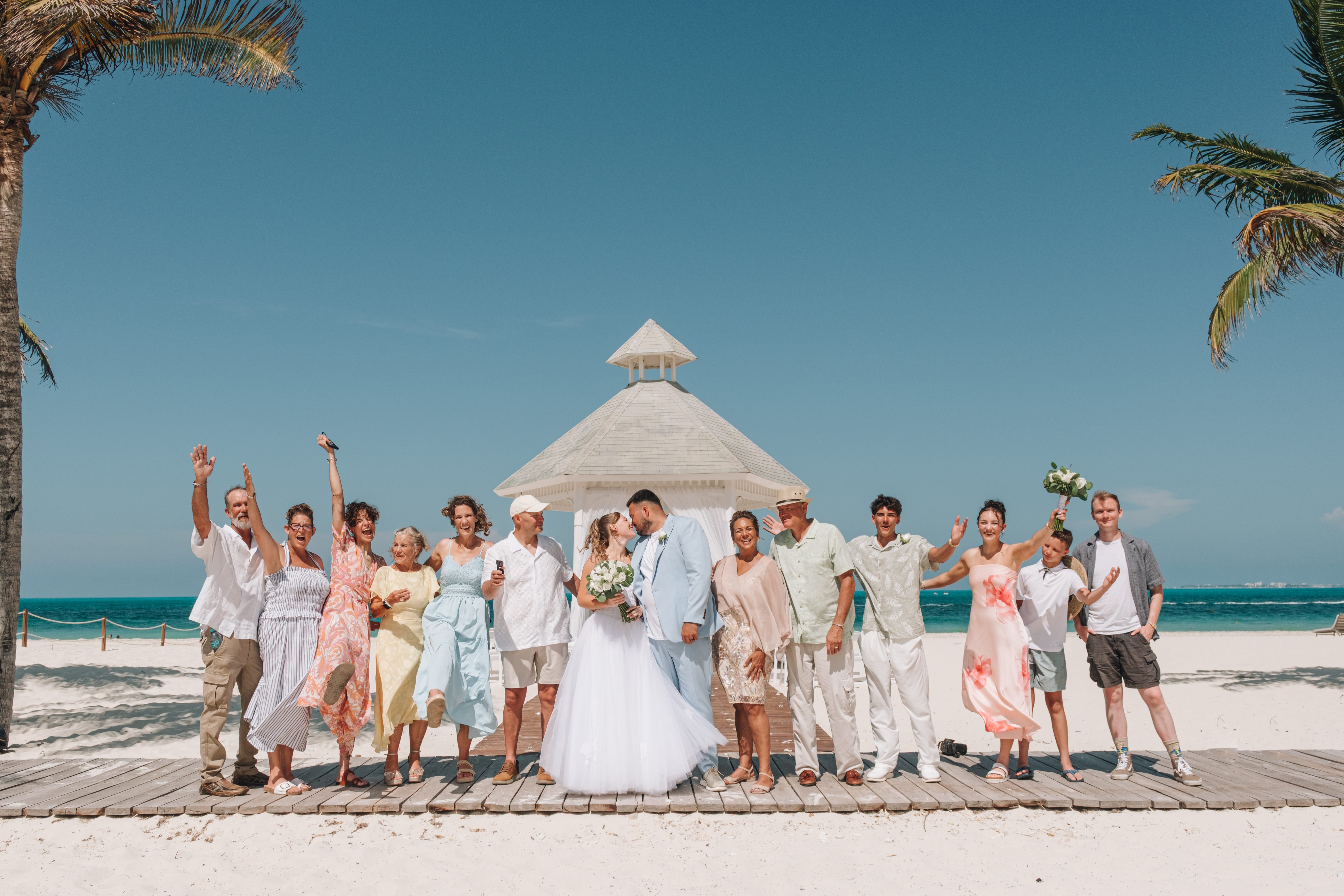 Kayleigh and Isaac share a kiss at their destination wedding, loved ones in summer outfits nearby.