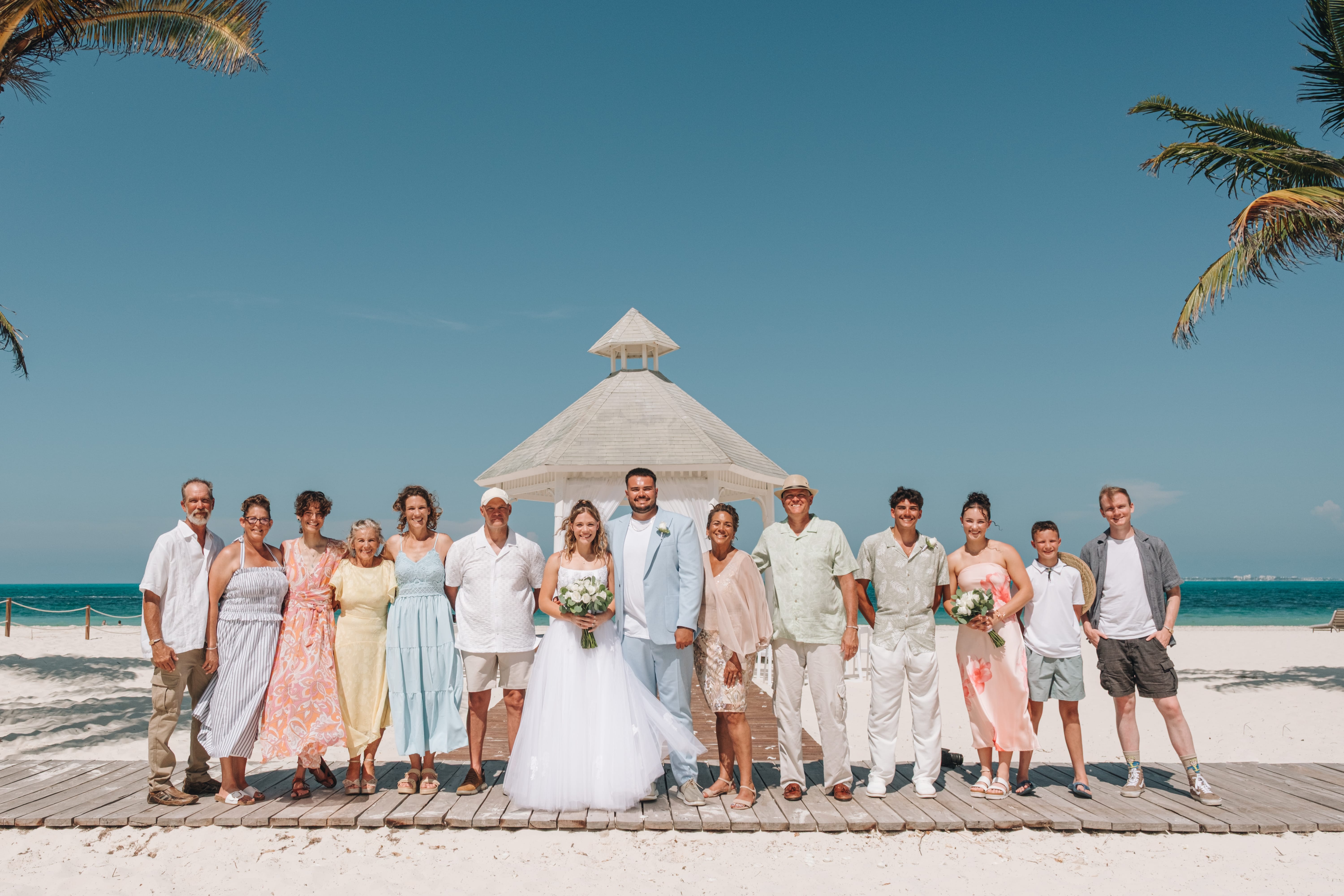 The destination wedding party stands on a boardwalk by a gazebo beneath a bright blue sky.