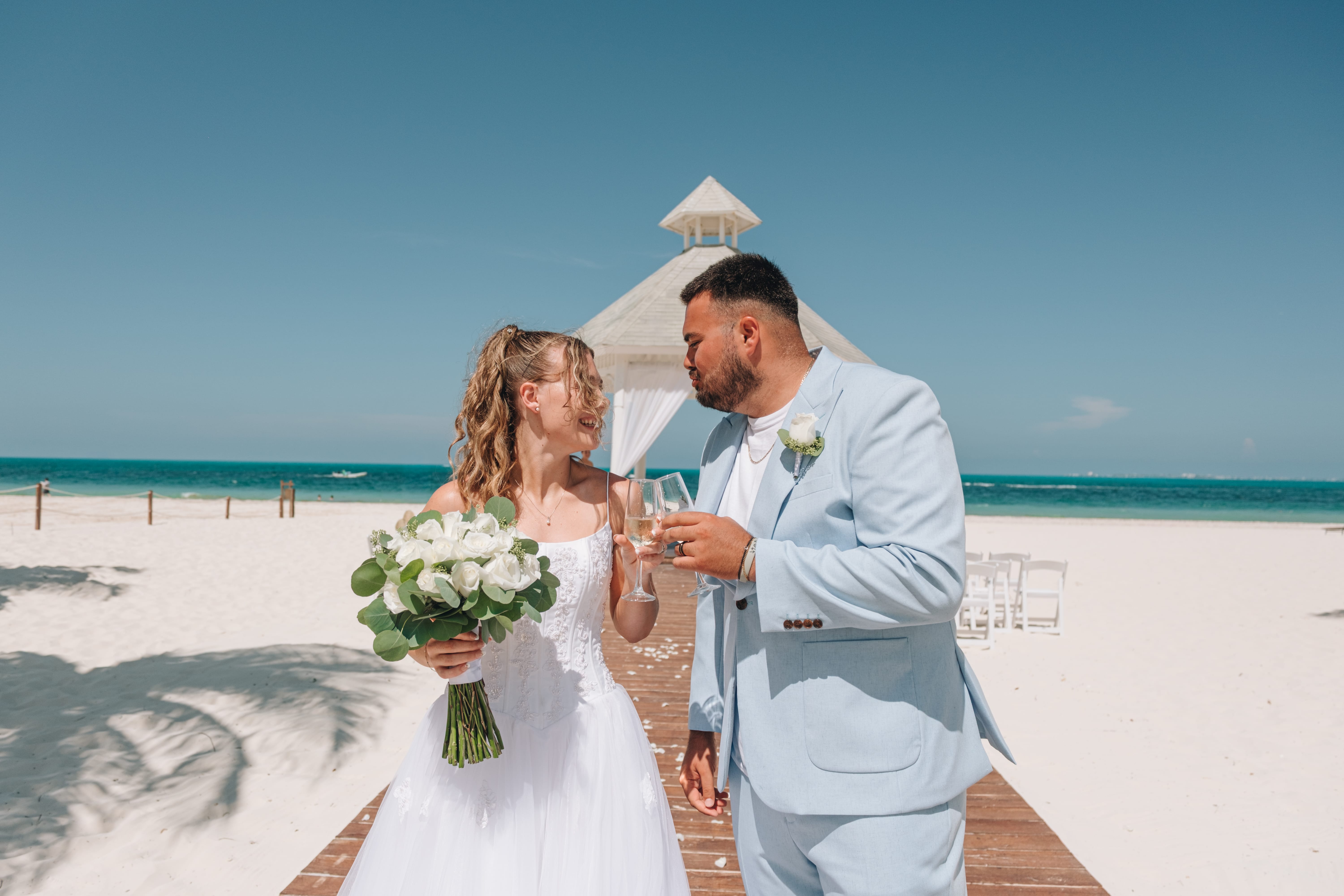 Kayleigh and Isaac toast at their destination wedding on a sandy beach, bouquet and waves behind.