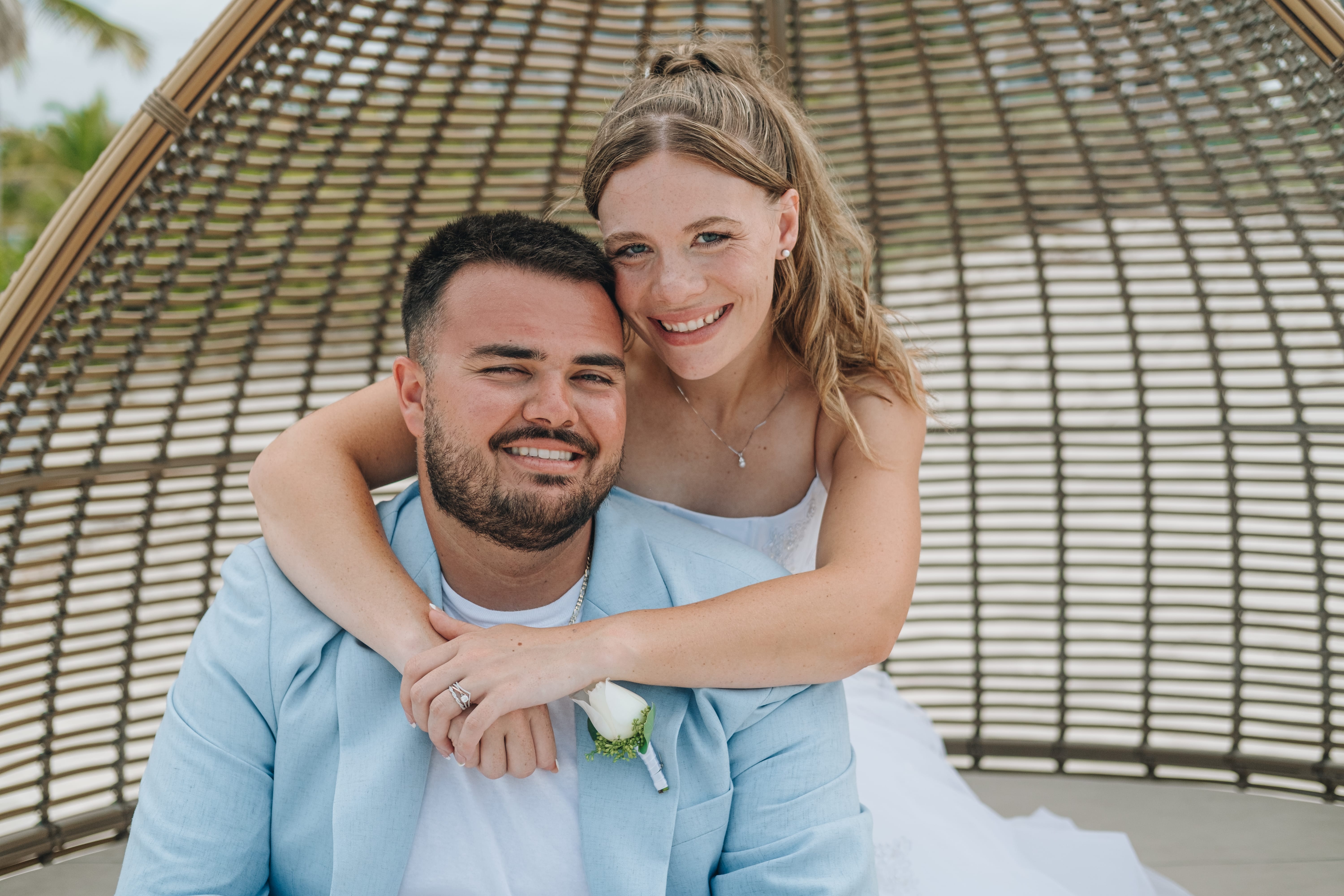 Kayleigh and Isaac smile on the beach under a wicker canopy at their destination wedding.