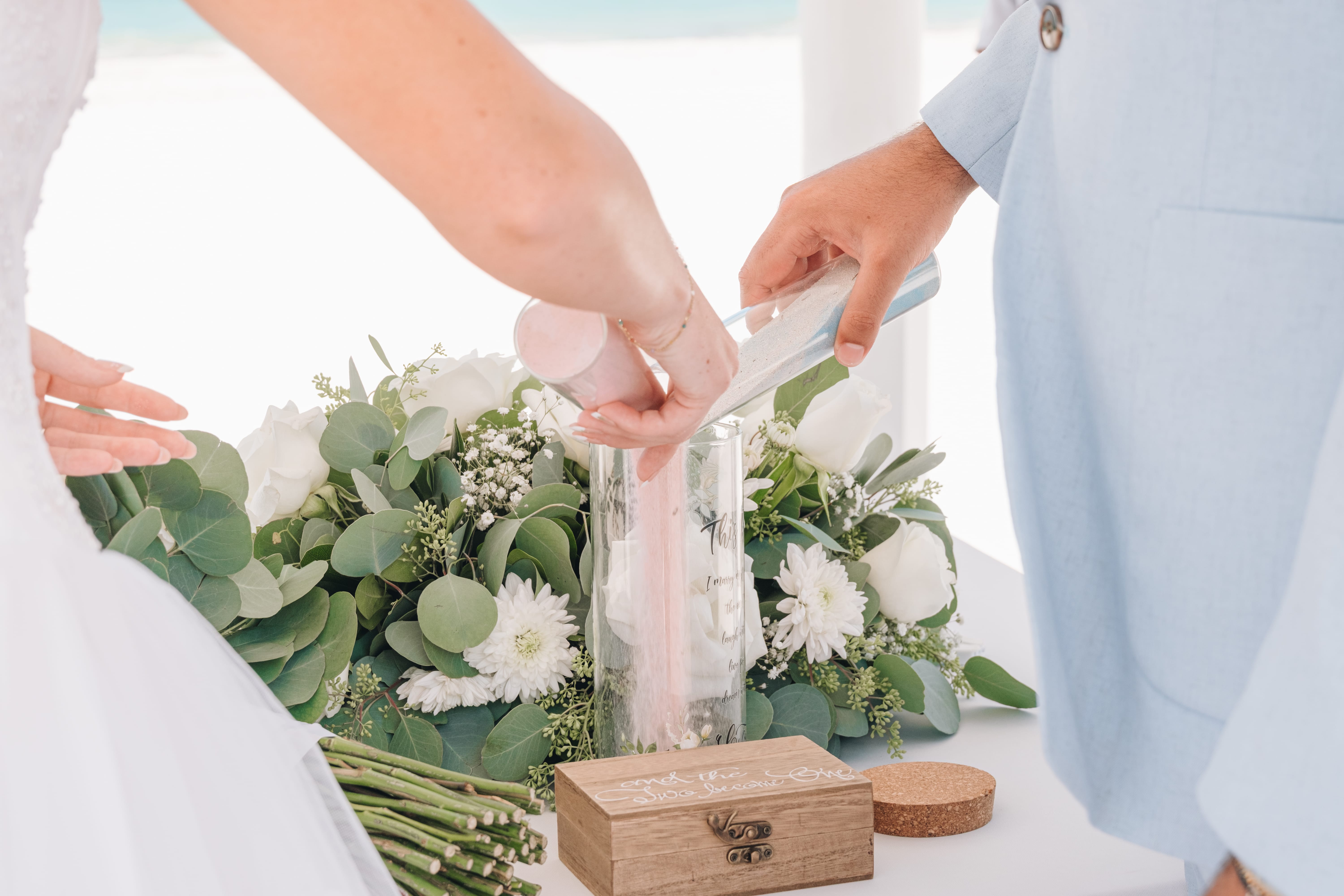 Sand from Kayleigh and Isaac is poured into a vase at a destination wedding near white flowers.