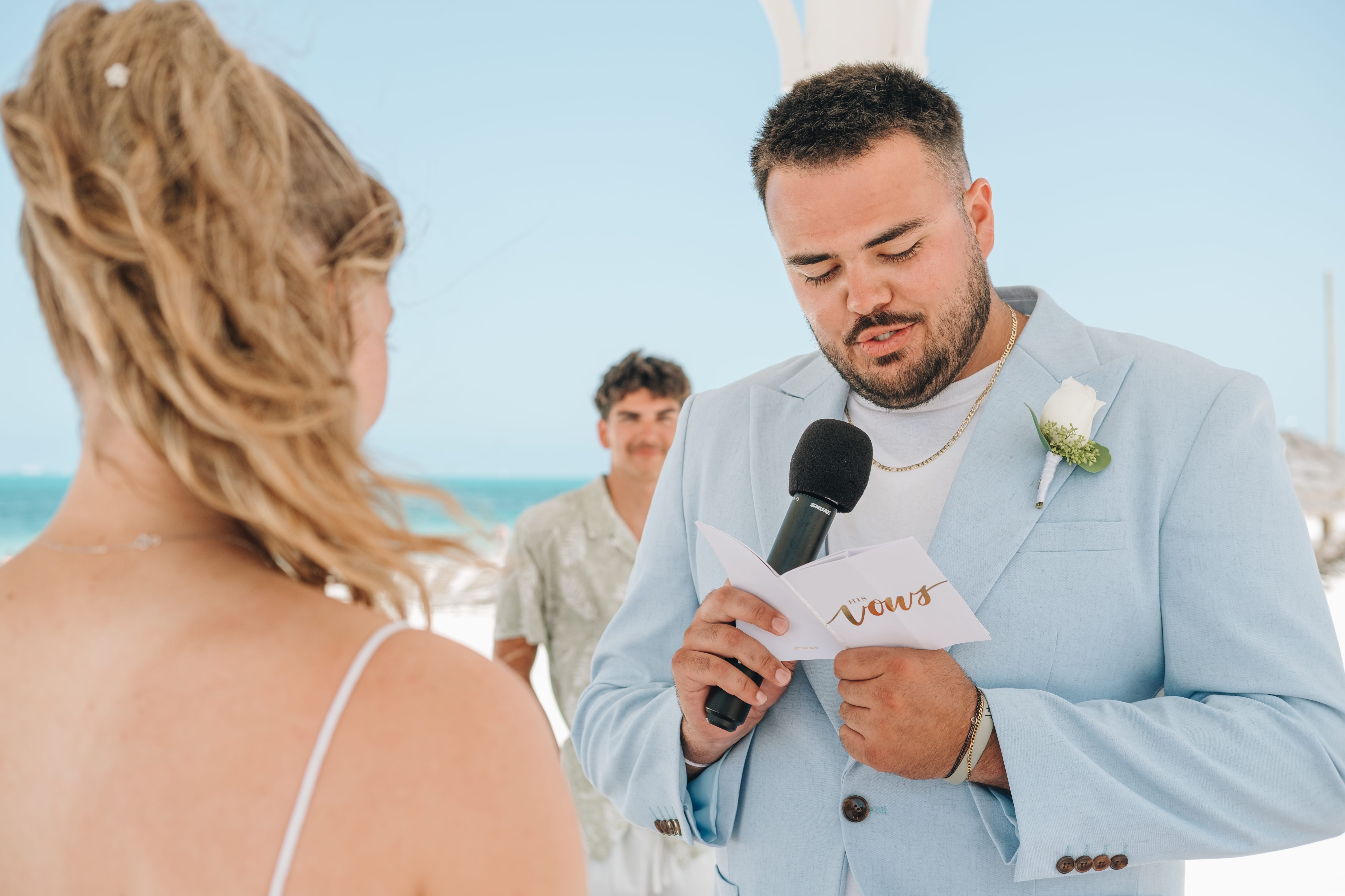 Destination wedding: groom in blue suit reads vows on the beach, officiant stands behind.