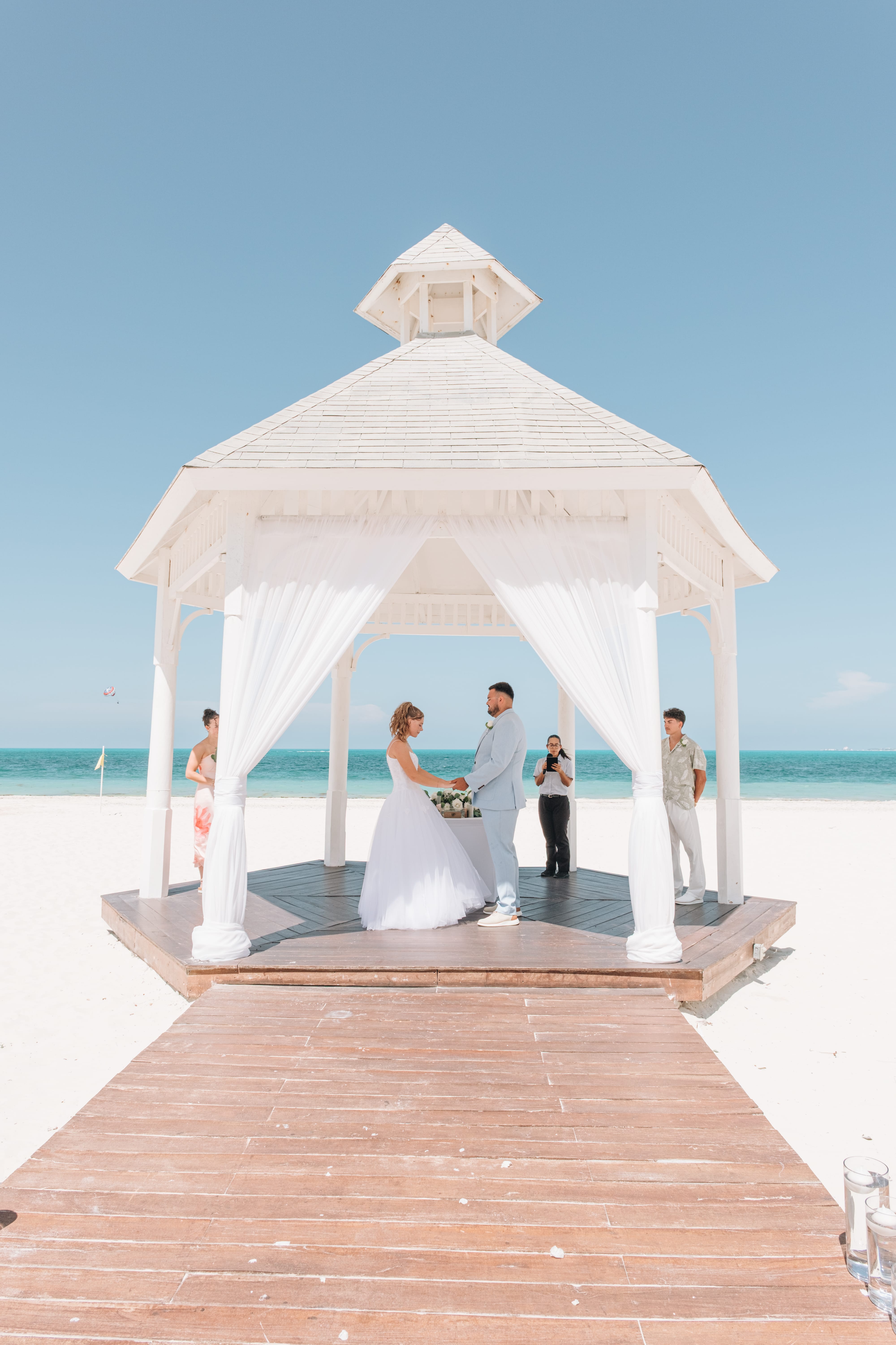 Kayleigh and Isaac exchange vows in a white gazebo at their destination wedding, loved ones nearby.