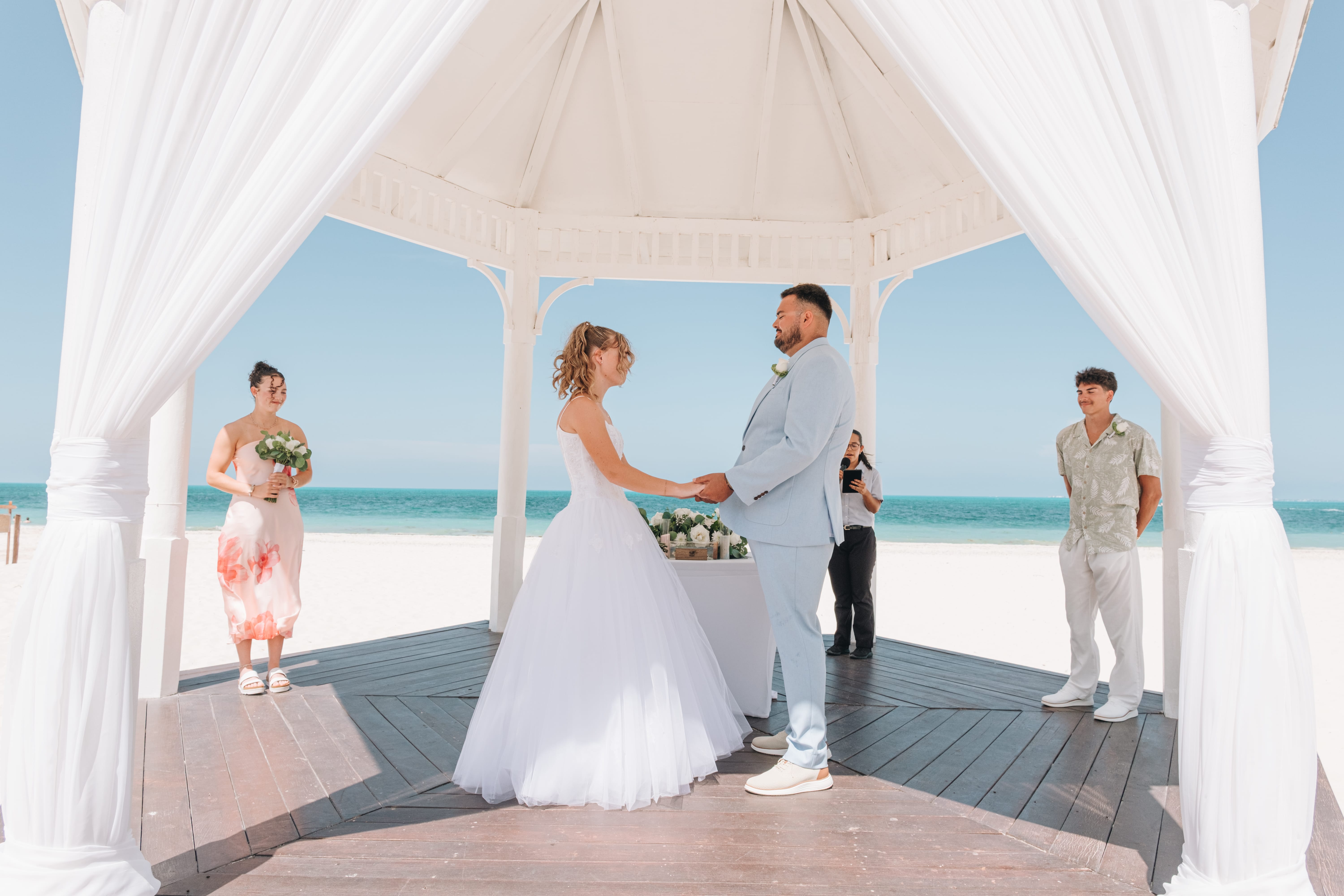 Kayleigh and Isaac wed under a white gazebo, holding hands, with three guests nearby.