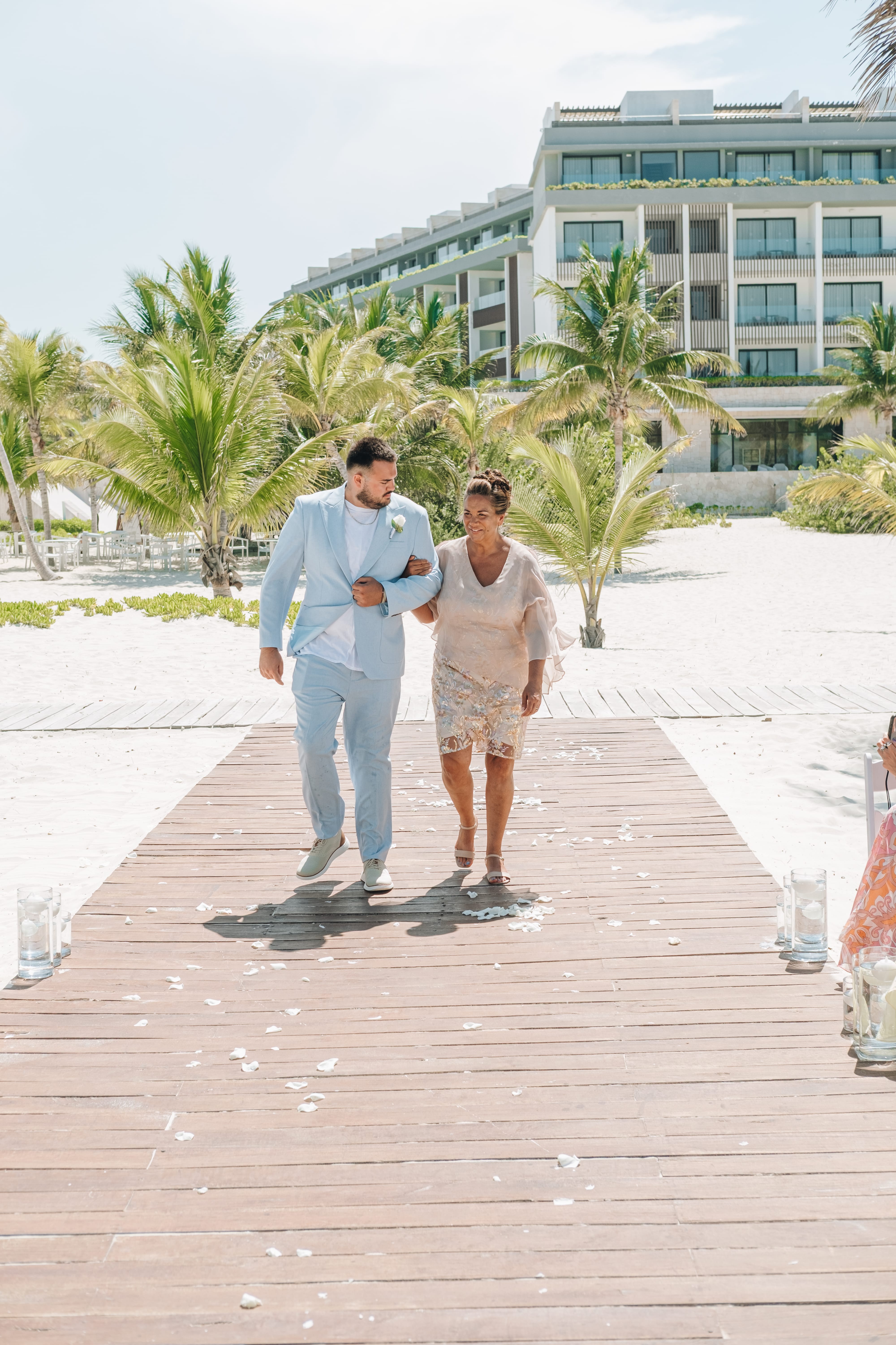 Kayleigh and Isaac walk arm in arm on a wooden path at their destination wedding beach resort.