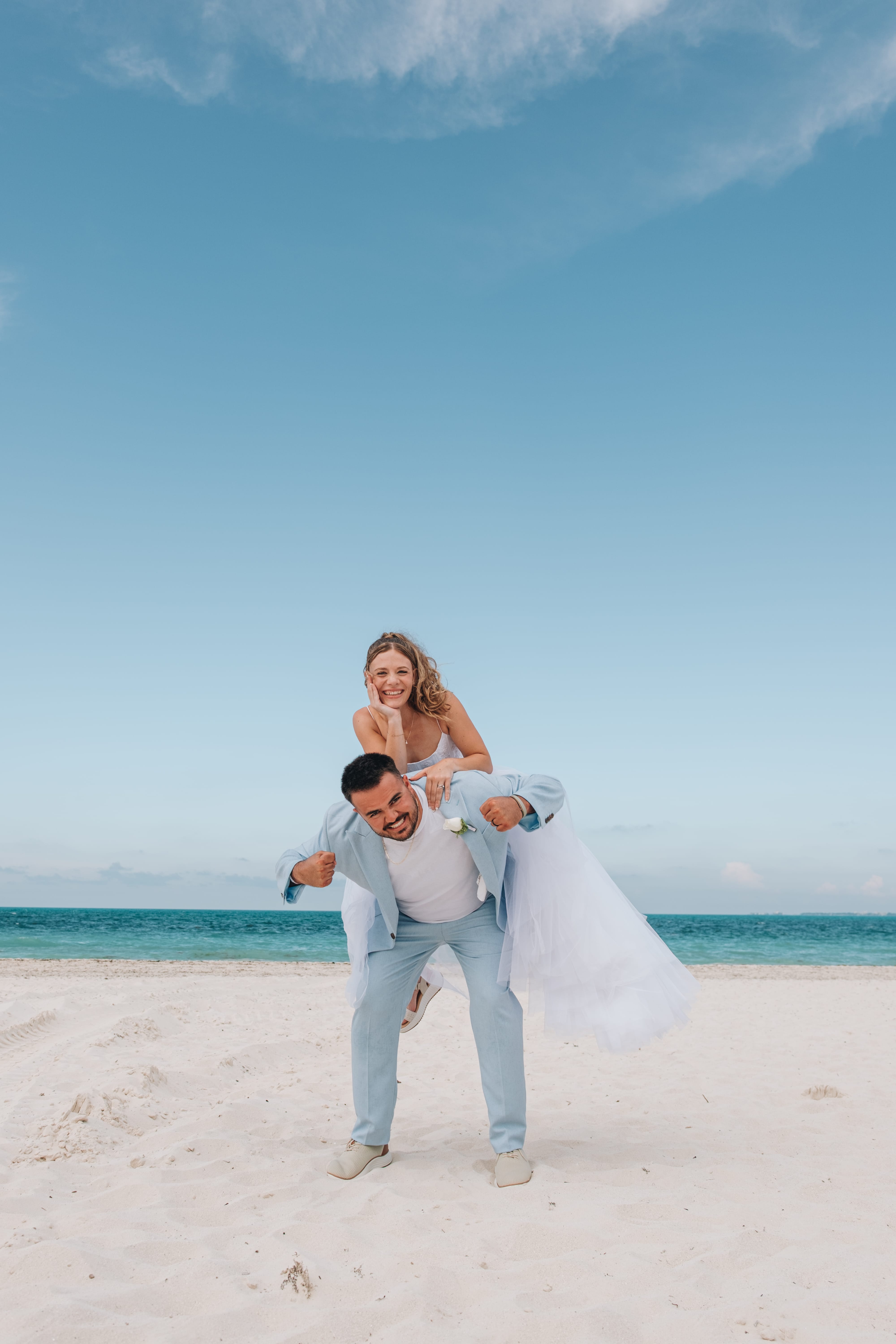 At their destination wedding, man in blue suit gives woman a piggyback ride on sandy beach.