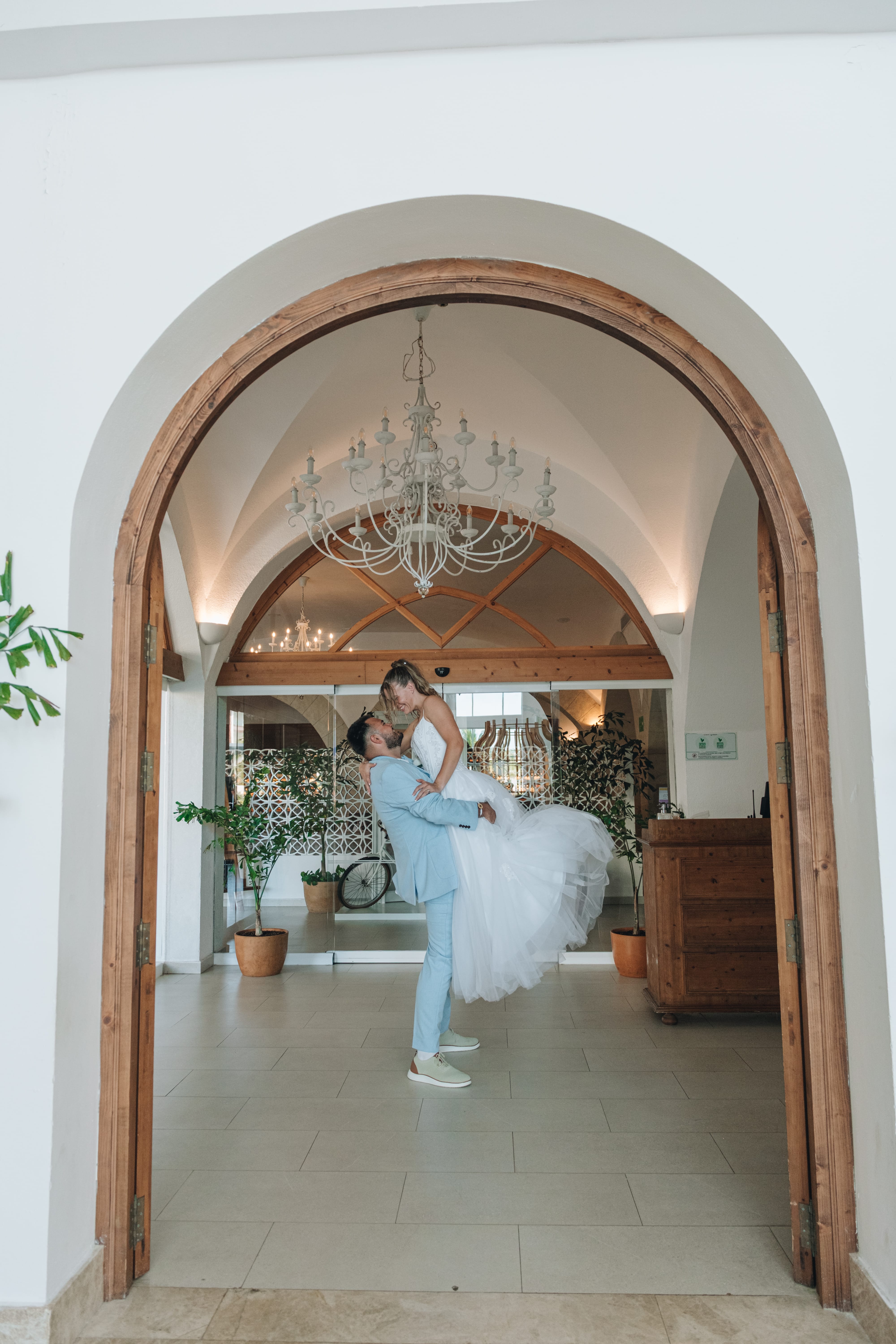 Kayleigh and Isaac: Groom in blue lifts bride in white at their destination wedding indoors.
