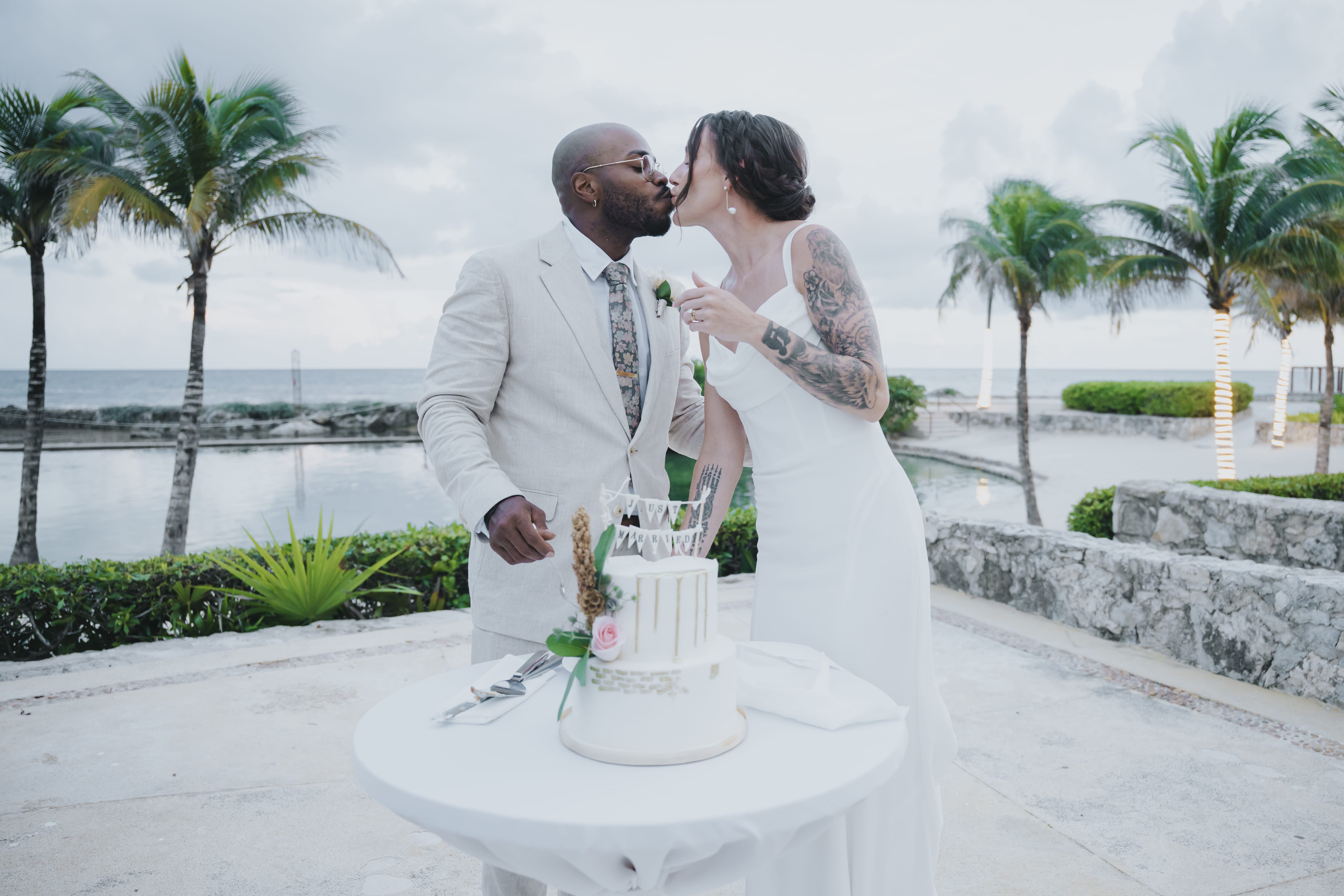 Madison and Blake share a kiss by their wedding cake at a destination wedding near palms and water.