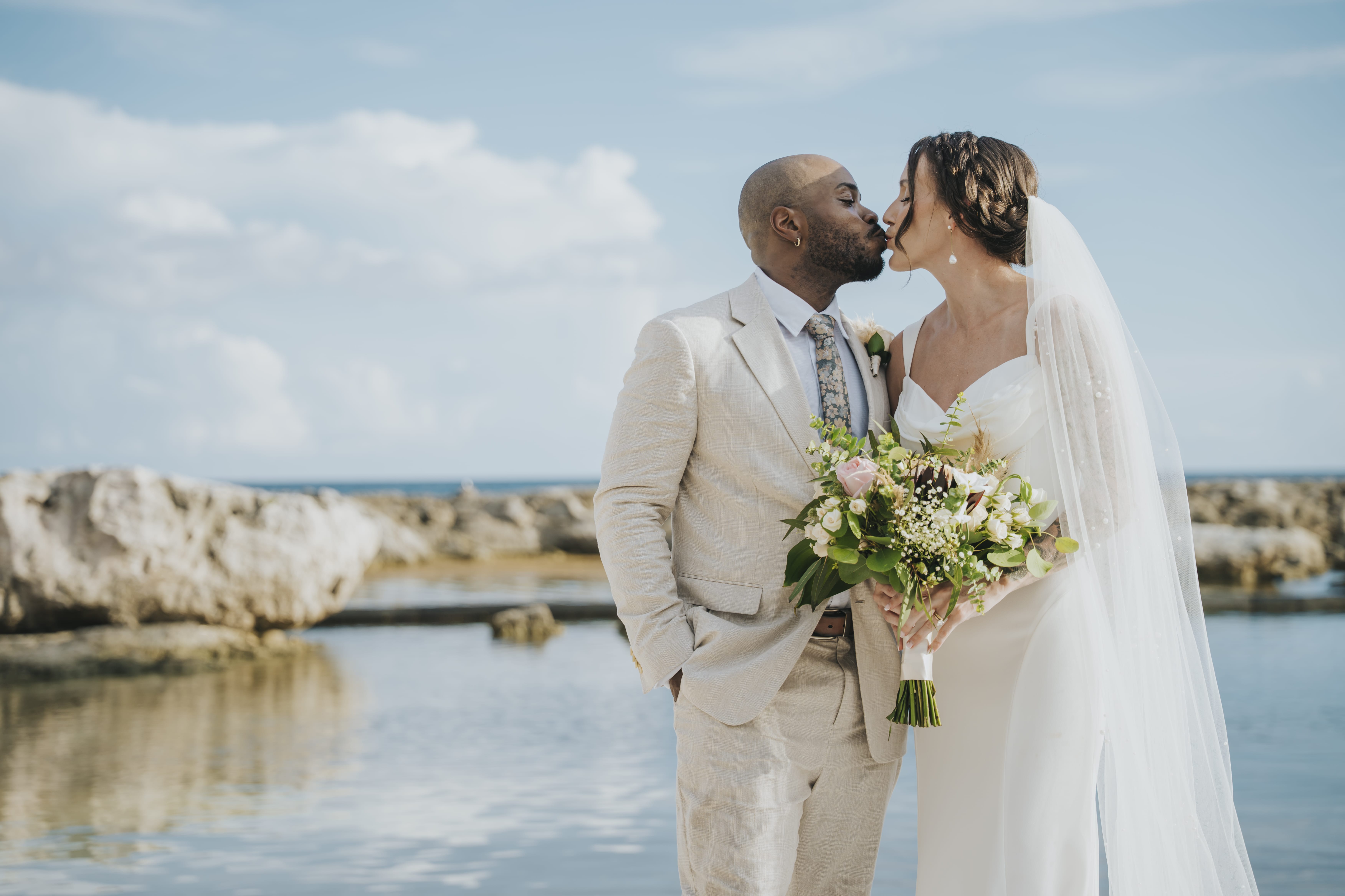 Madison and Blake share a kiss by water at their destination wedding, bride holds bouquet.