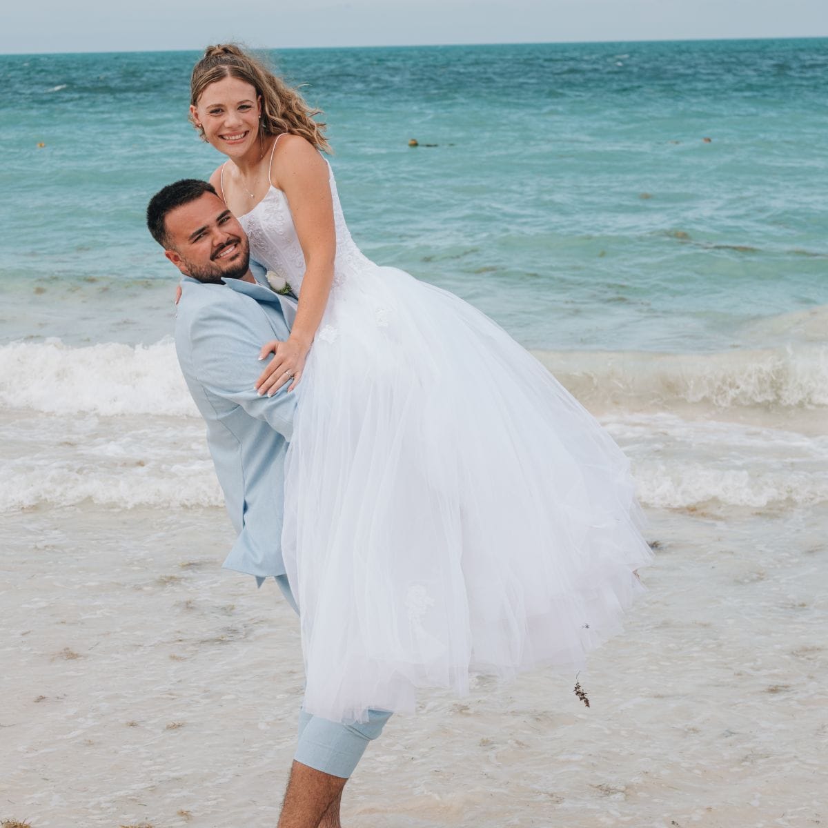 At a destination wedding, groom in light suit lifts bride in white gown on beach with waves behind.