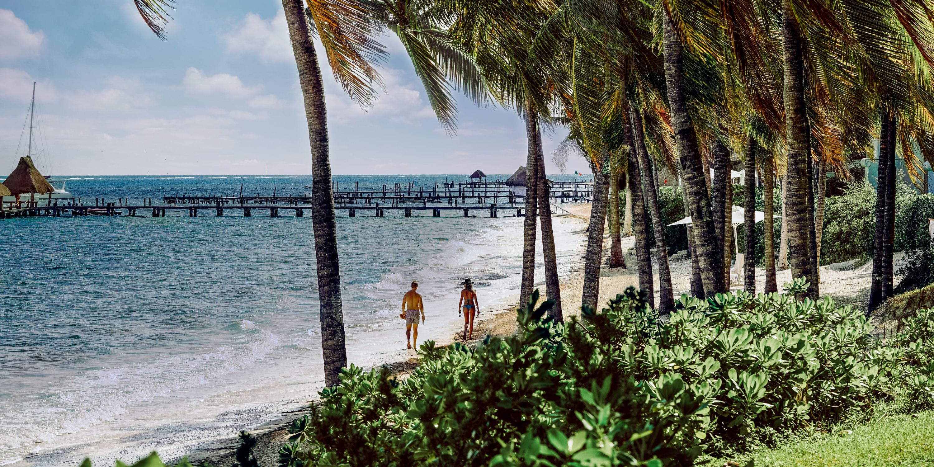 A couple walks along a beach with palm trees at a Royalton Riviera Cancun destination wedding.