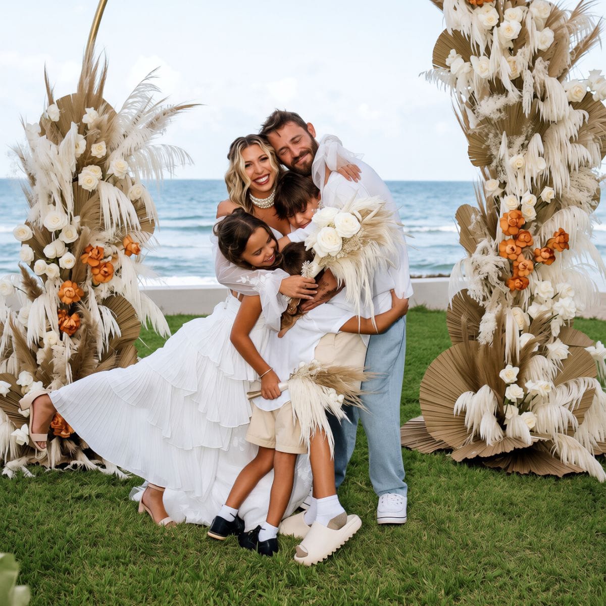 A family in white hugs by a floral beach arch at a destination wedding. Product: Kayleigh and Isaac.