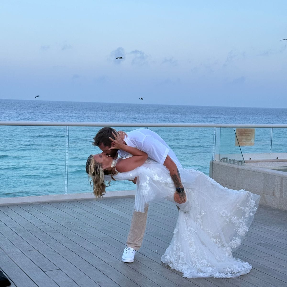 A destination wedding couple shares a kiss on a sunset-lit deck overlooking the ocean.