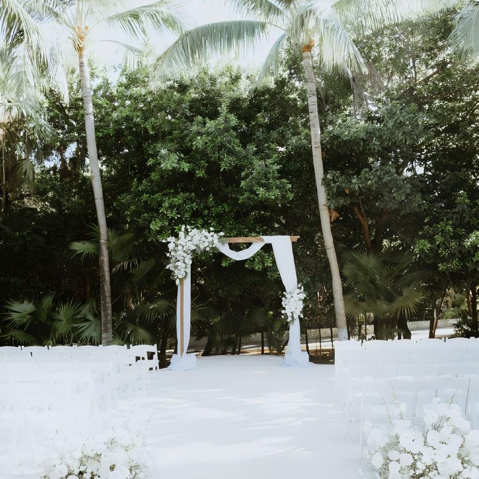 Destination wedding arch adorned with flowers, set outdoors among palm trees and white chairs.