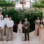 Destination wedding party in formal attire poses on outdoor steps, greenery backdrop behind them.