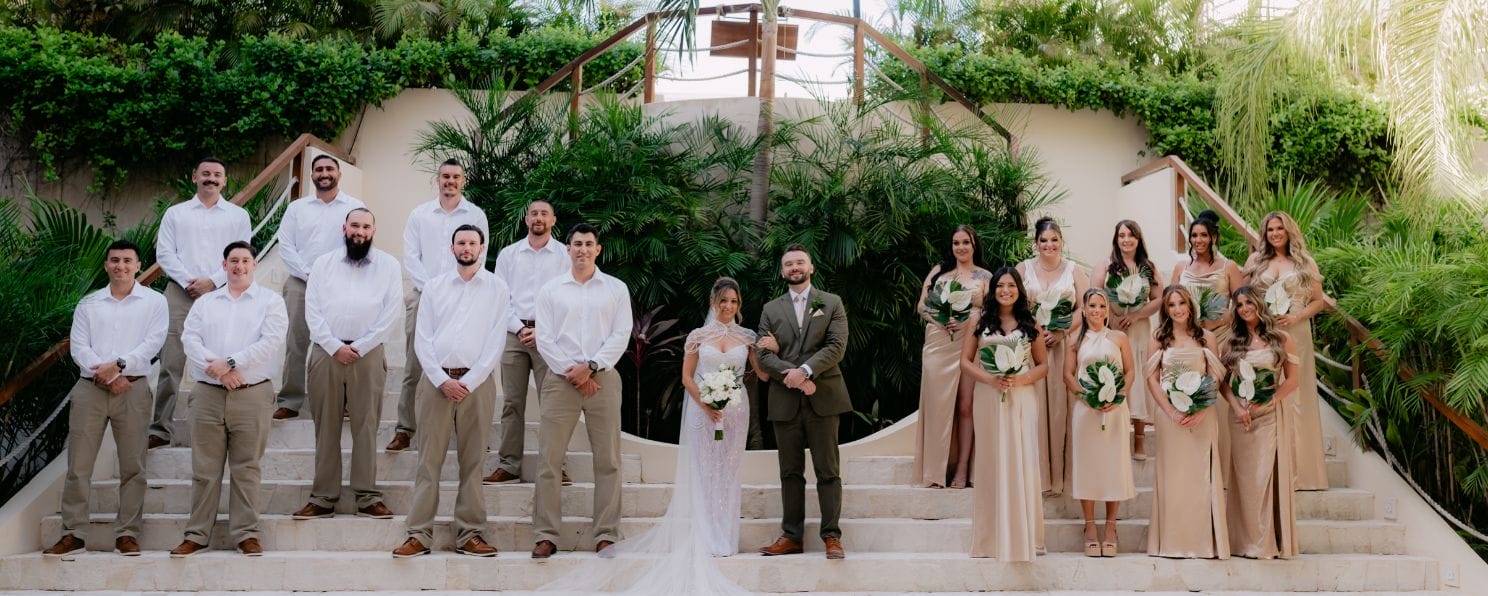 Destination wedding party in formal attire poses on outdoor steps, greenery backdrop behind them.