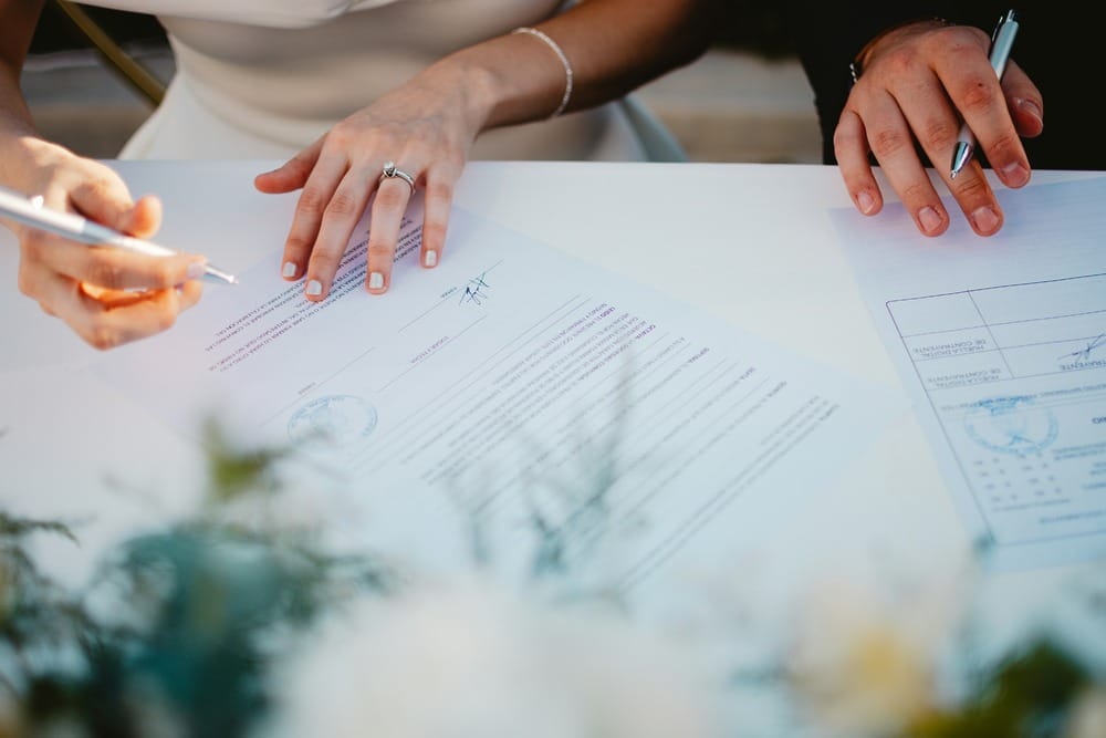 Hands sign wedding documents at a table; only the papers and hands are visible in the scene.