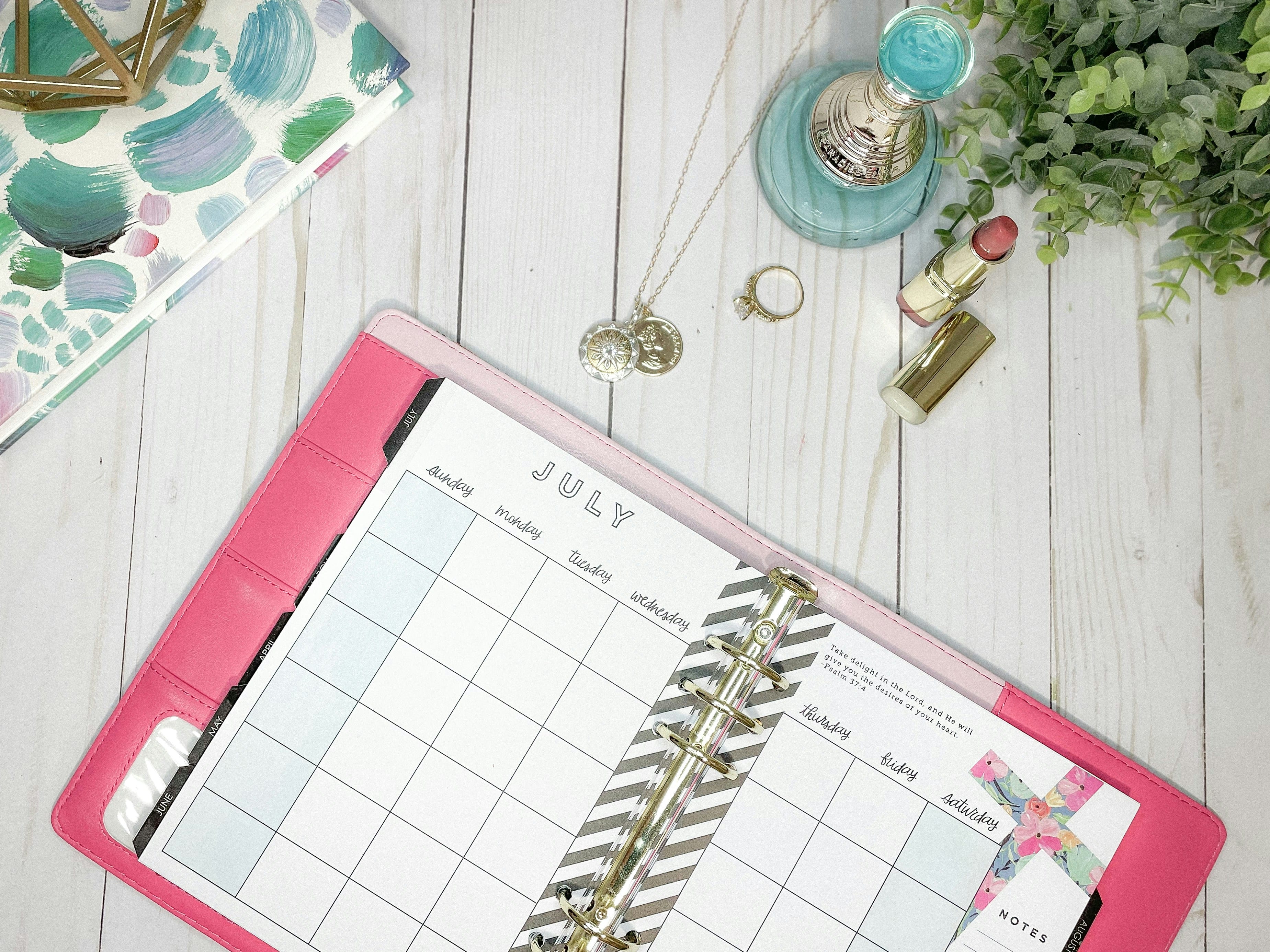 Open planner with July calendar, lipstick, jewelry, and plant on desk for destination wedding prep.