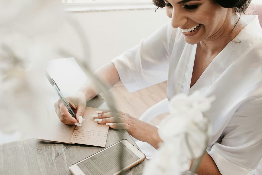 Bride in a white robe writes wedding notes at a table, her phone beside her.