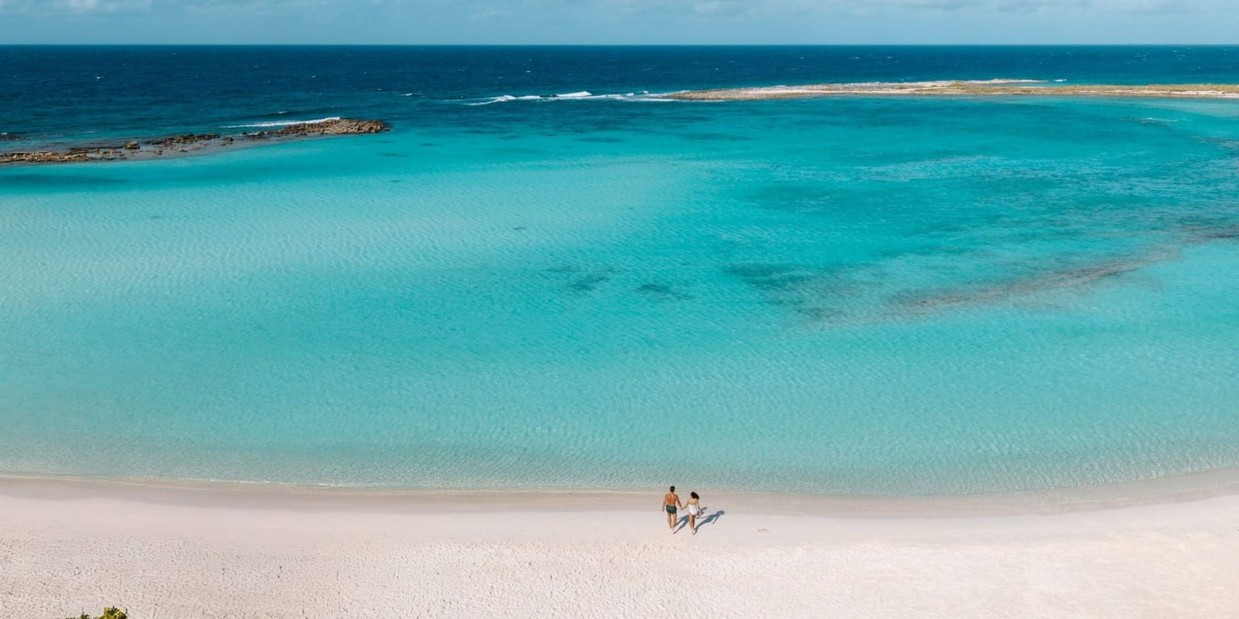 Three people stroll a sandy beach at a destination wedding, clear turquoise water and island nearby.