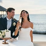 Newlyweds in formal wear toast champagne at a destination wedding cake table by the ocean.
