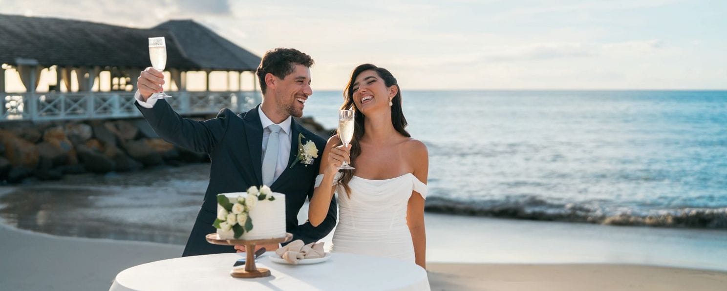 Newlyweds in formal wear toast champagne at a destination wedding cake table by the ocean.