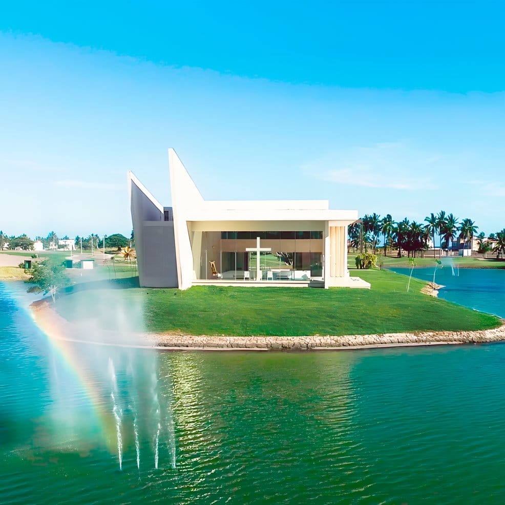 Modern white chapel on grassy island with fountains and rainbow; ideal for destination weddings.