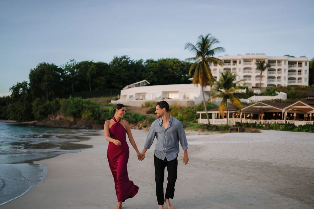 A couple walks barefoot on the beach at sunset, holding hands near a destination wedding resort.