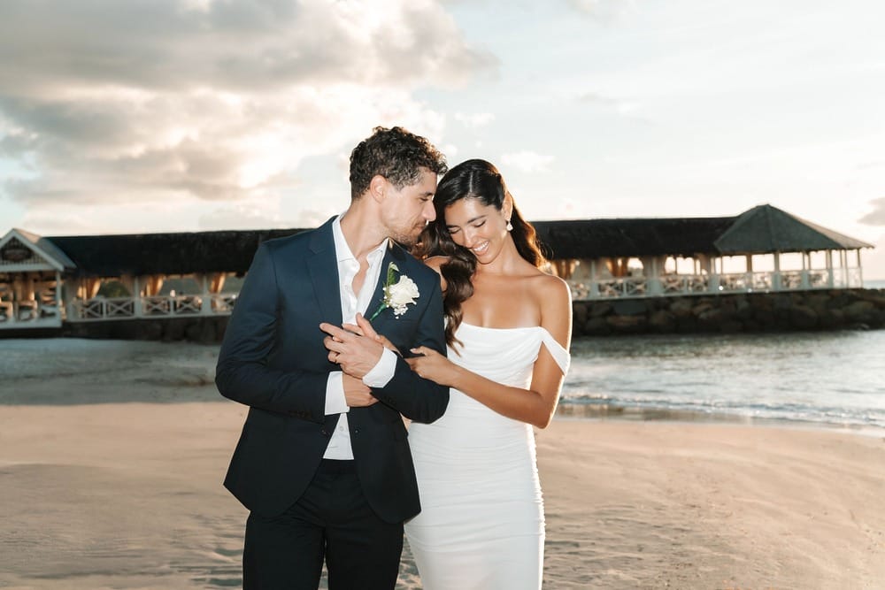 Bride and groom embrace on a sandy beach at sunset during their destination wedding near a pavilion.